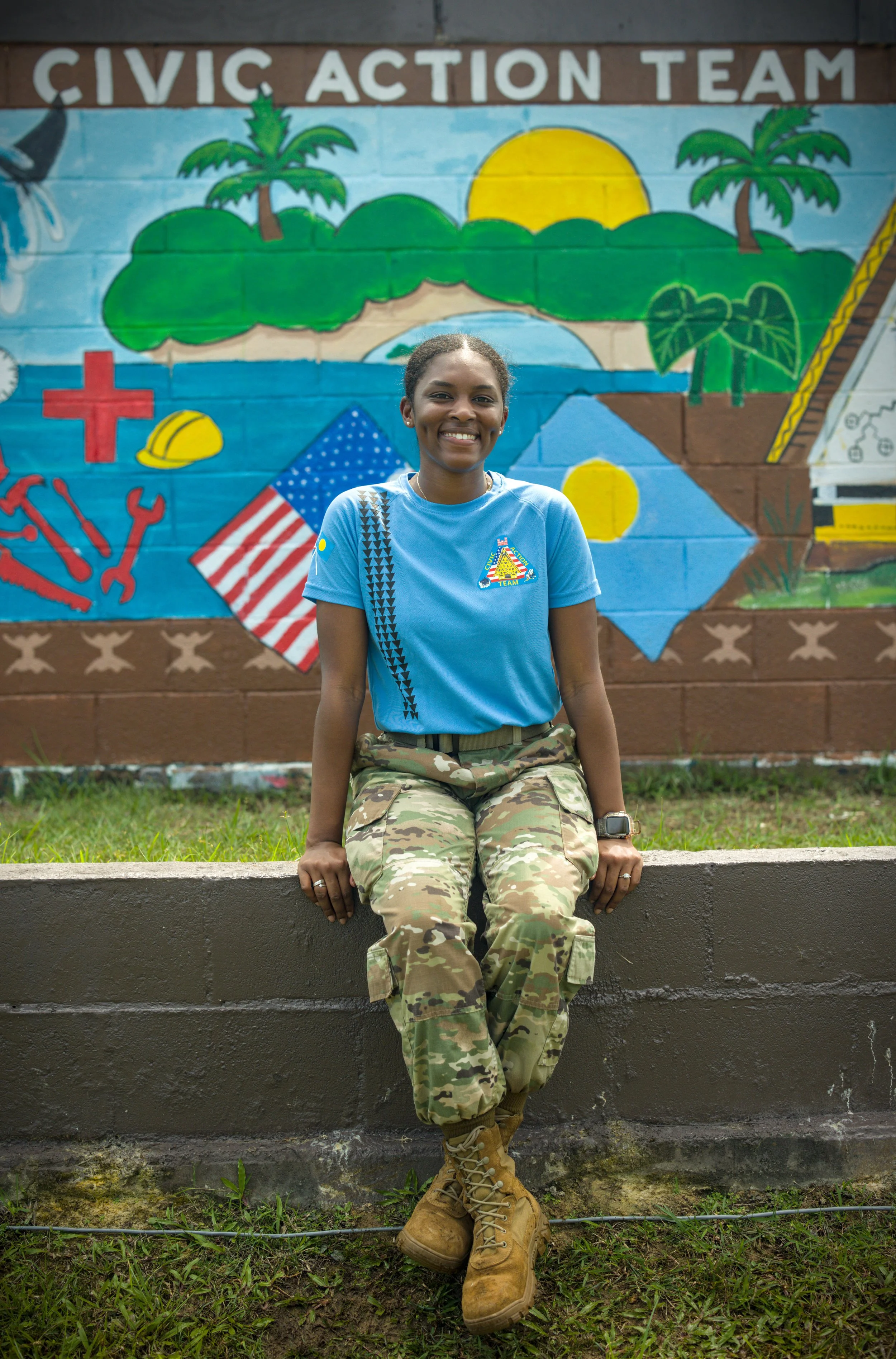 Young female soldier in camouflage uniform smiling, standing against a plain beige wall.