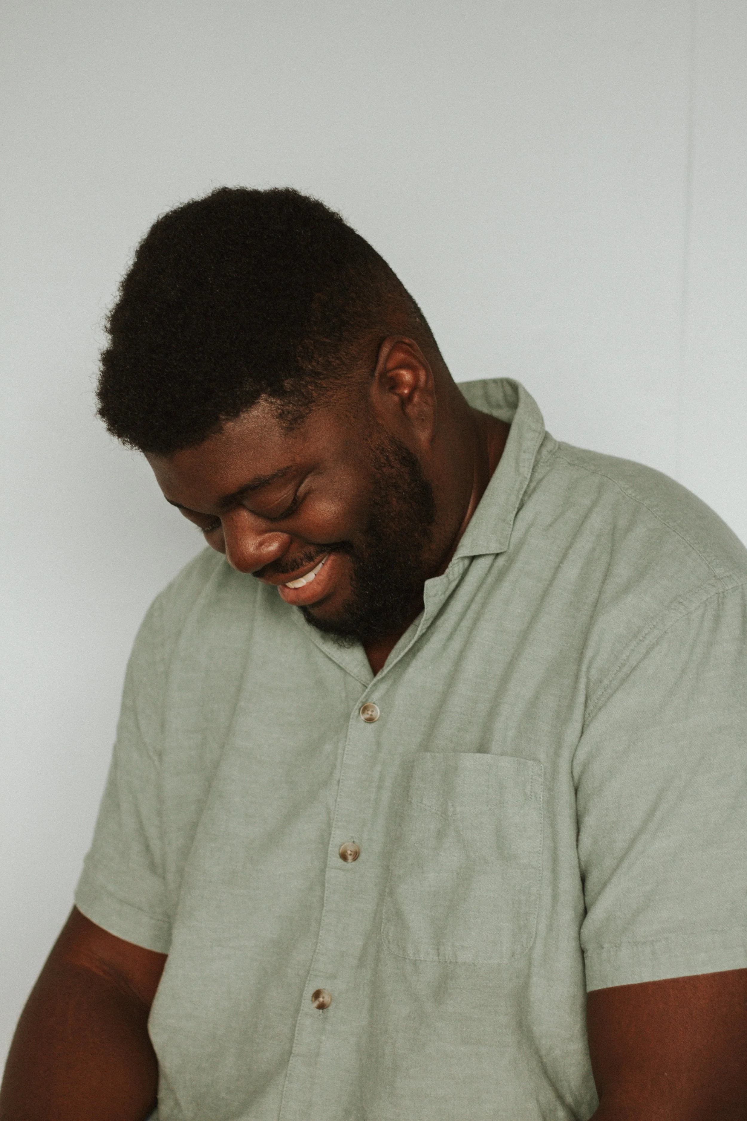 Smiling man with short curly hair and beard, wearing a light green button-down shirt, looking downward, against a plain white background.