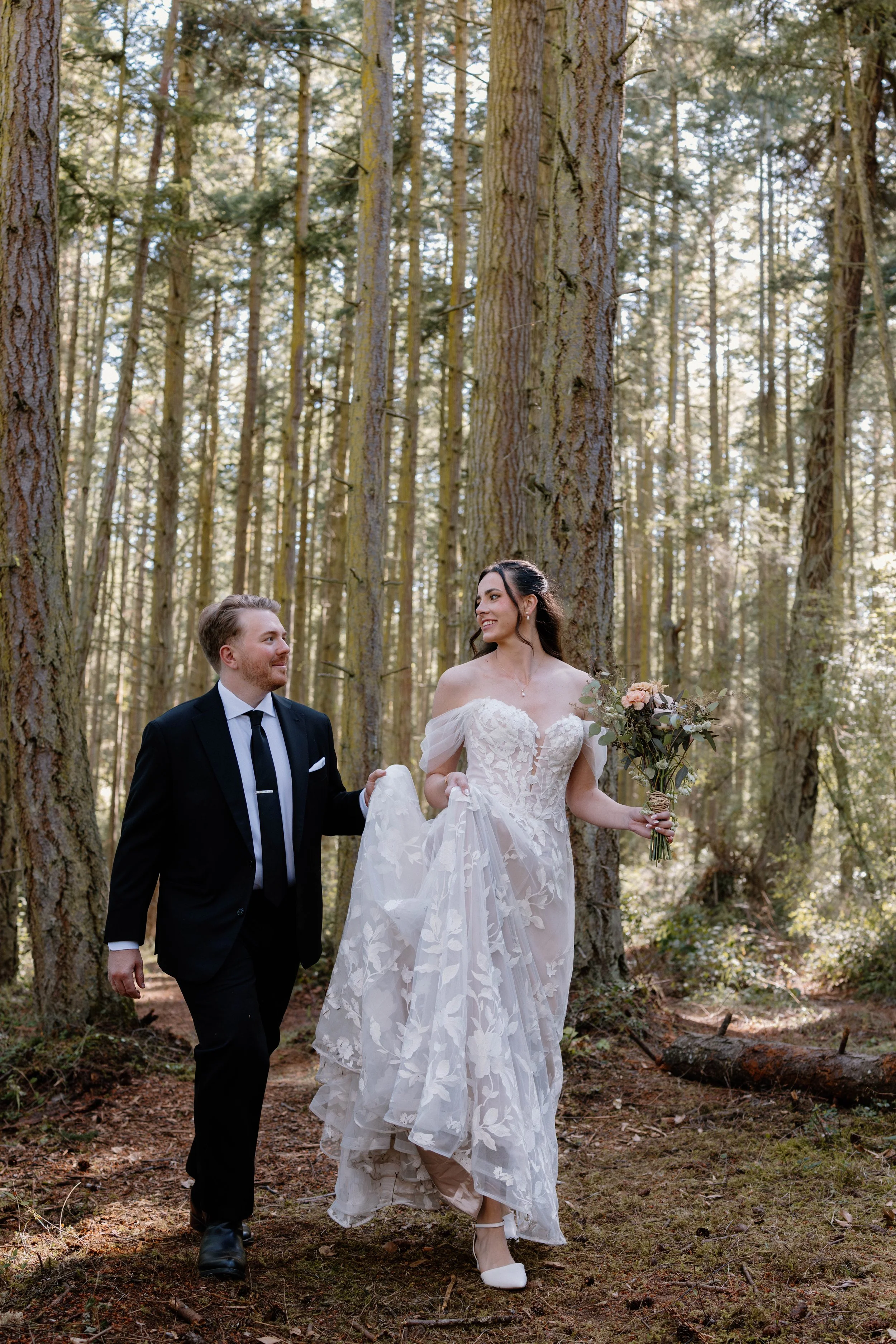 A bride and groom walking through a forest, holding hands, with tall trees and sunlight filtering through the branches.