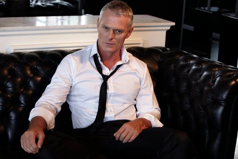A man with short blonde hair wearing a white dress shirt and a black tie, sitting on a black leather tufted sofa, looking at the camera with a serious expression.