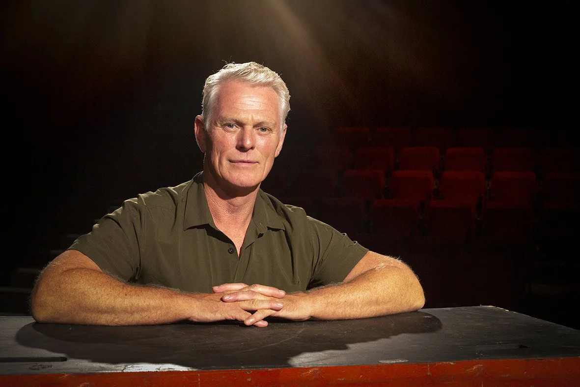 Older man with gray hair sitting at a table in a dimly lit auditorium or theater.