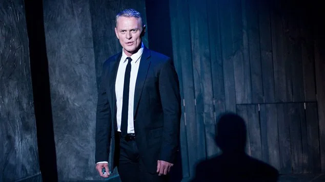 A man in a black suit and tie standing in front of a dark background with textured wall and wooden panels, with his shadow visible on the wall.