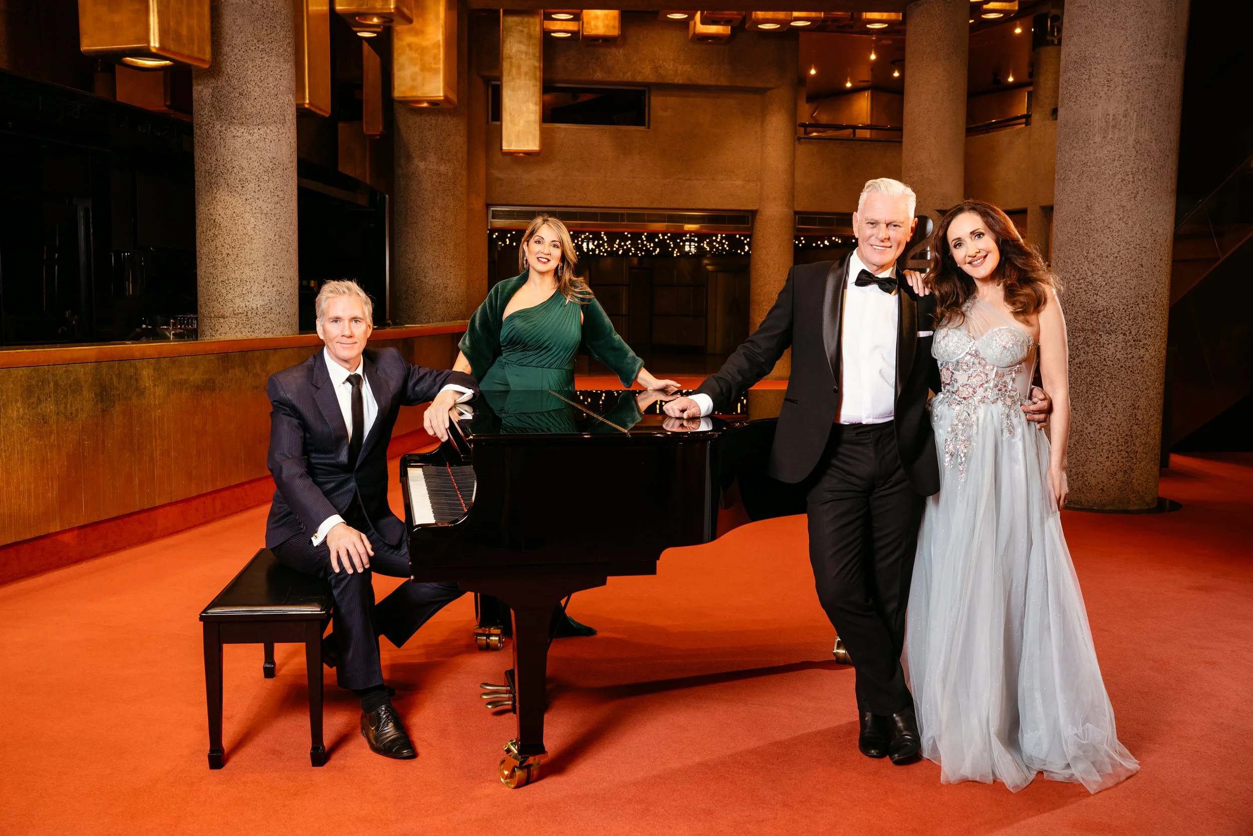 Four people in formal attire posing around a grand piano in a warmly lit, elegant venue. Two women and two men, with the women in evening gowns and the men in tuxedos, smiling at the camera.