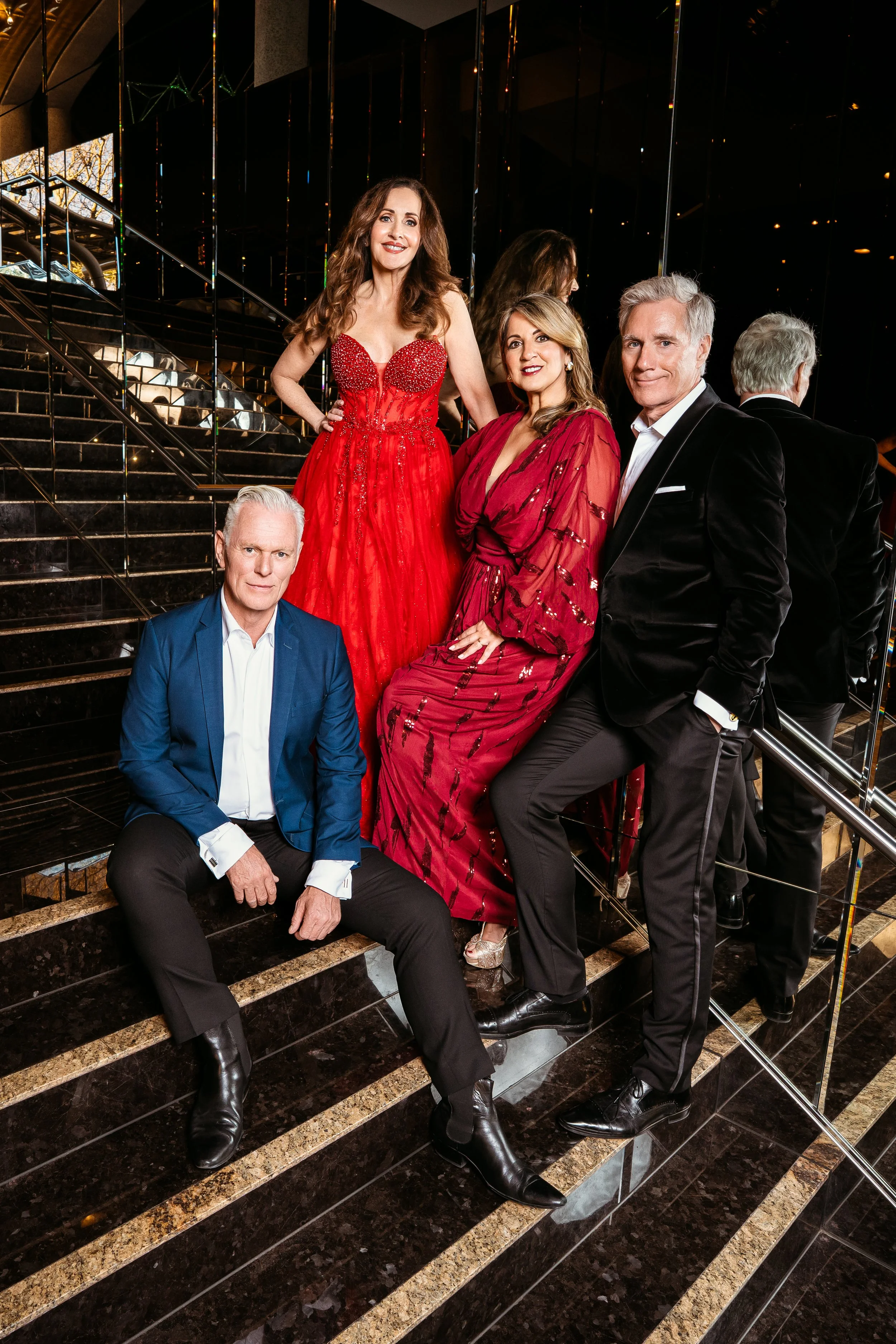Five well-dressed people posing on a black staircase with reflective surfaces and glass railings, with some people mirrored in the background.