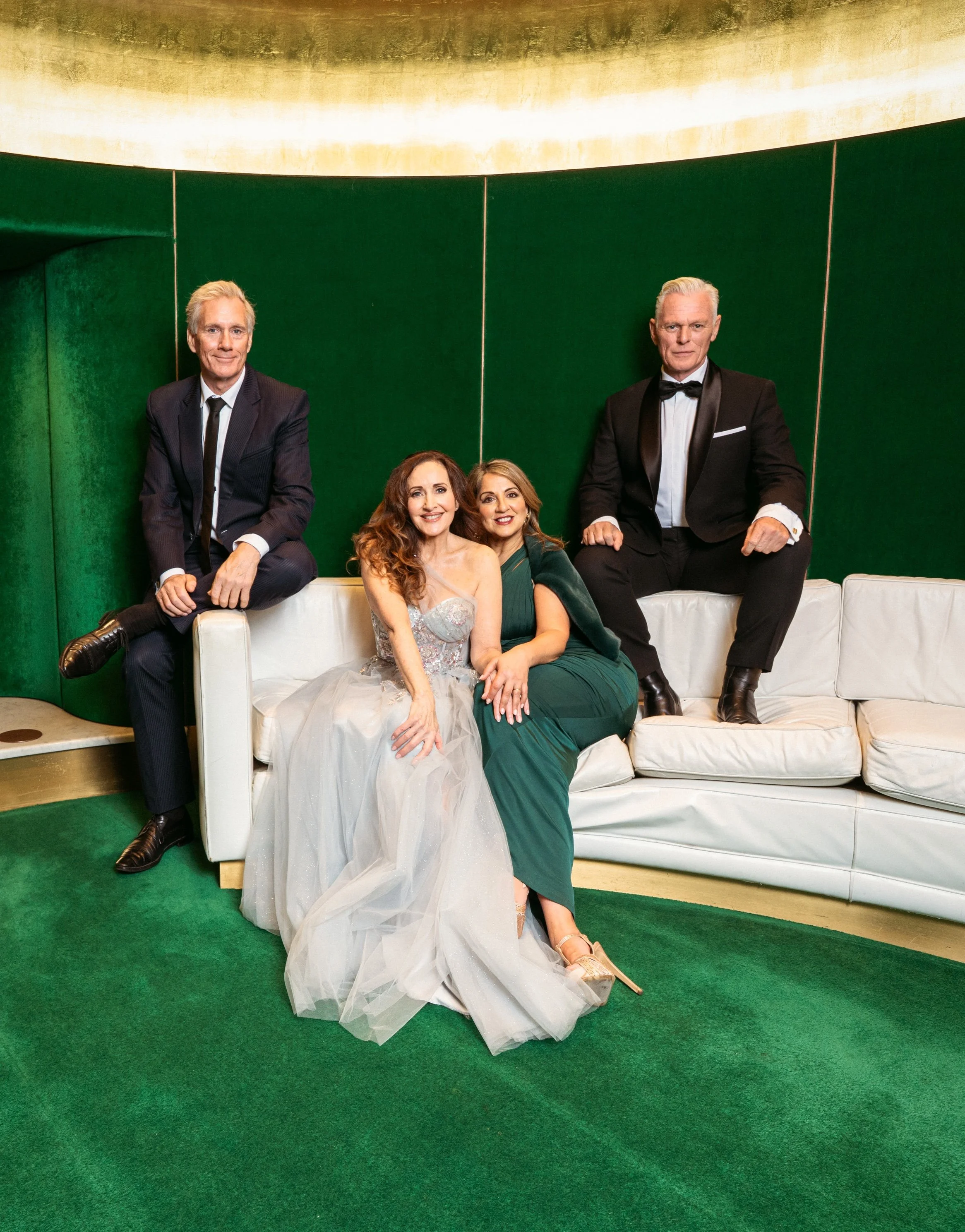 Four well-dressed individuals, two men in tuxedos and two women, are sitting on and around a white couch in an elegant setting with green walls and gold accents.