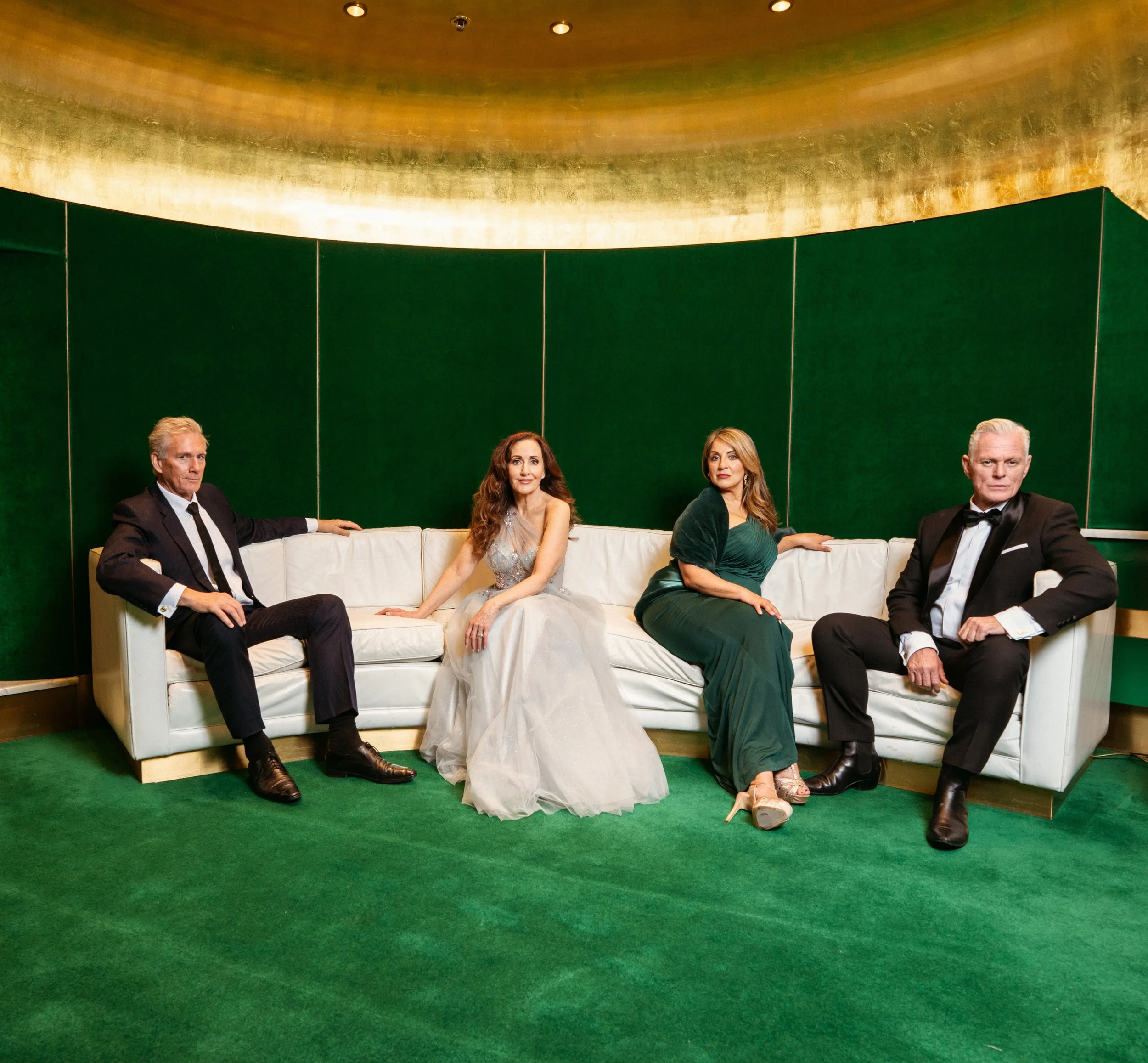 Four people sitting on a white sofa in a room with green walls and a gold ceiling. Two men in tuxedos and two women in evening dresses.