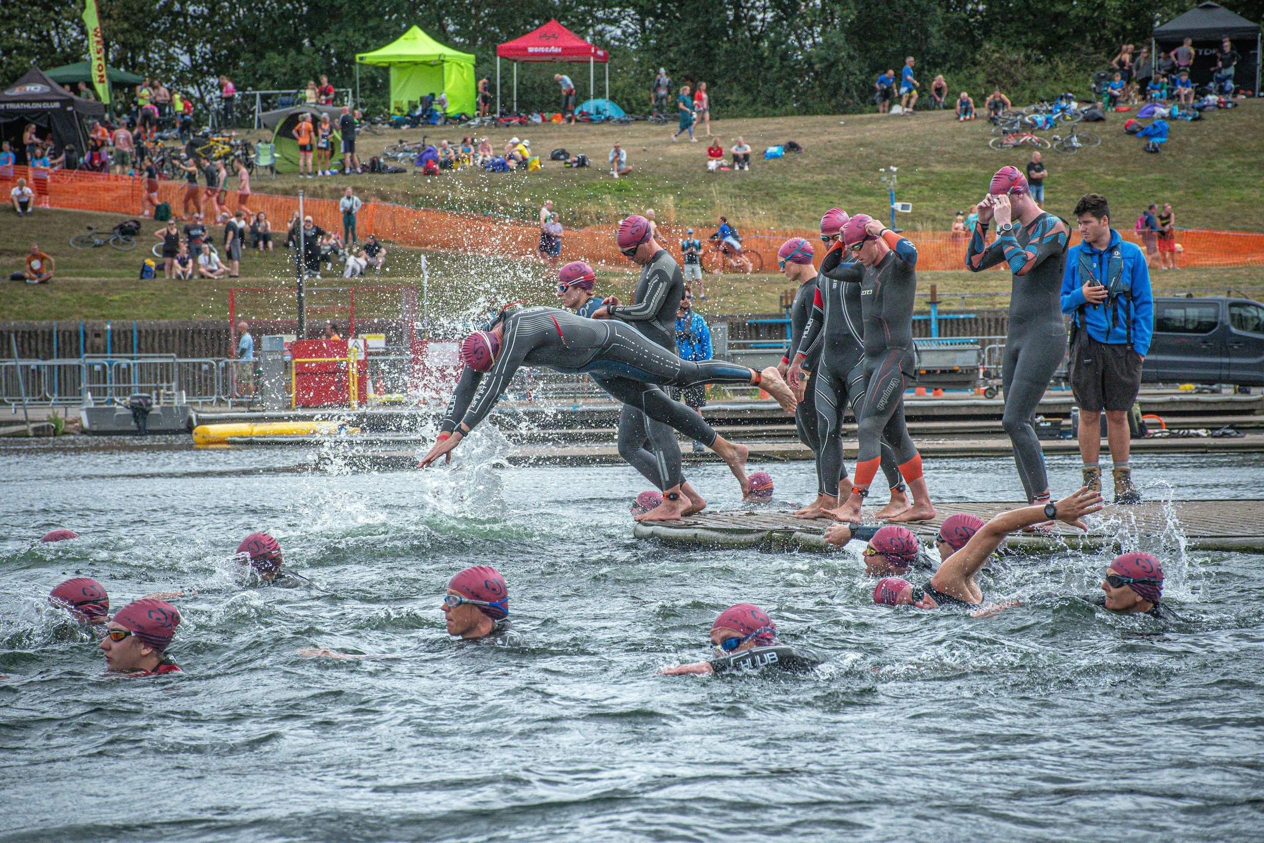 Swimmers with pink caps diving into water during a triathlon event, with spectators and tents on a grassy area in the background.