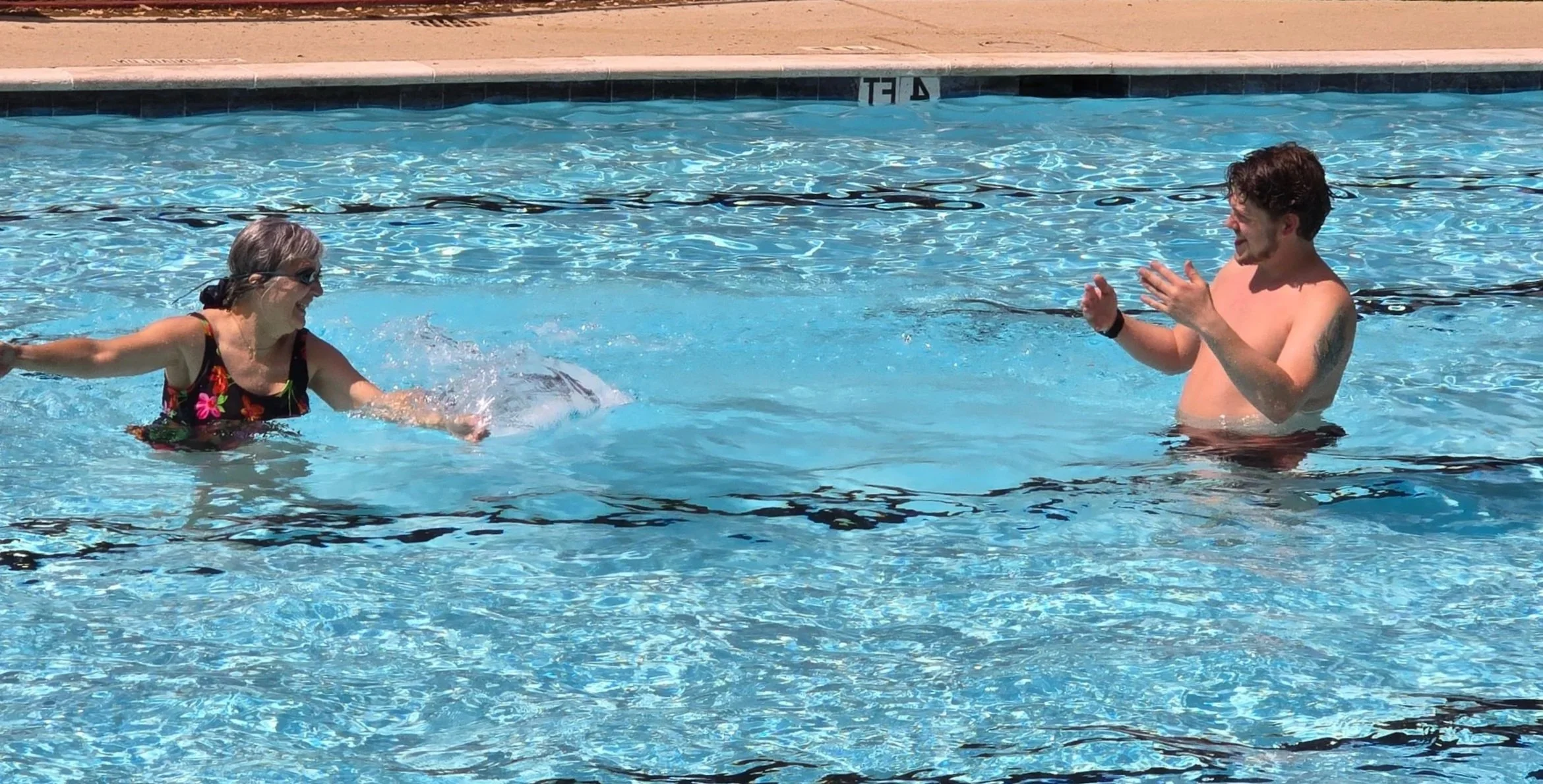 A woman and a man playing and having fun in a swimming pool on a sunny day. The woman is wearing a floral swimsuit and sunglasses, while the man is shirtless and clapping his hands.