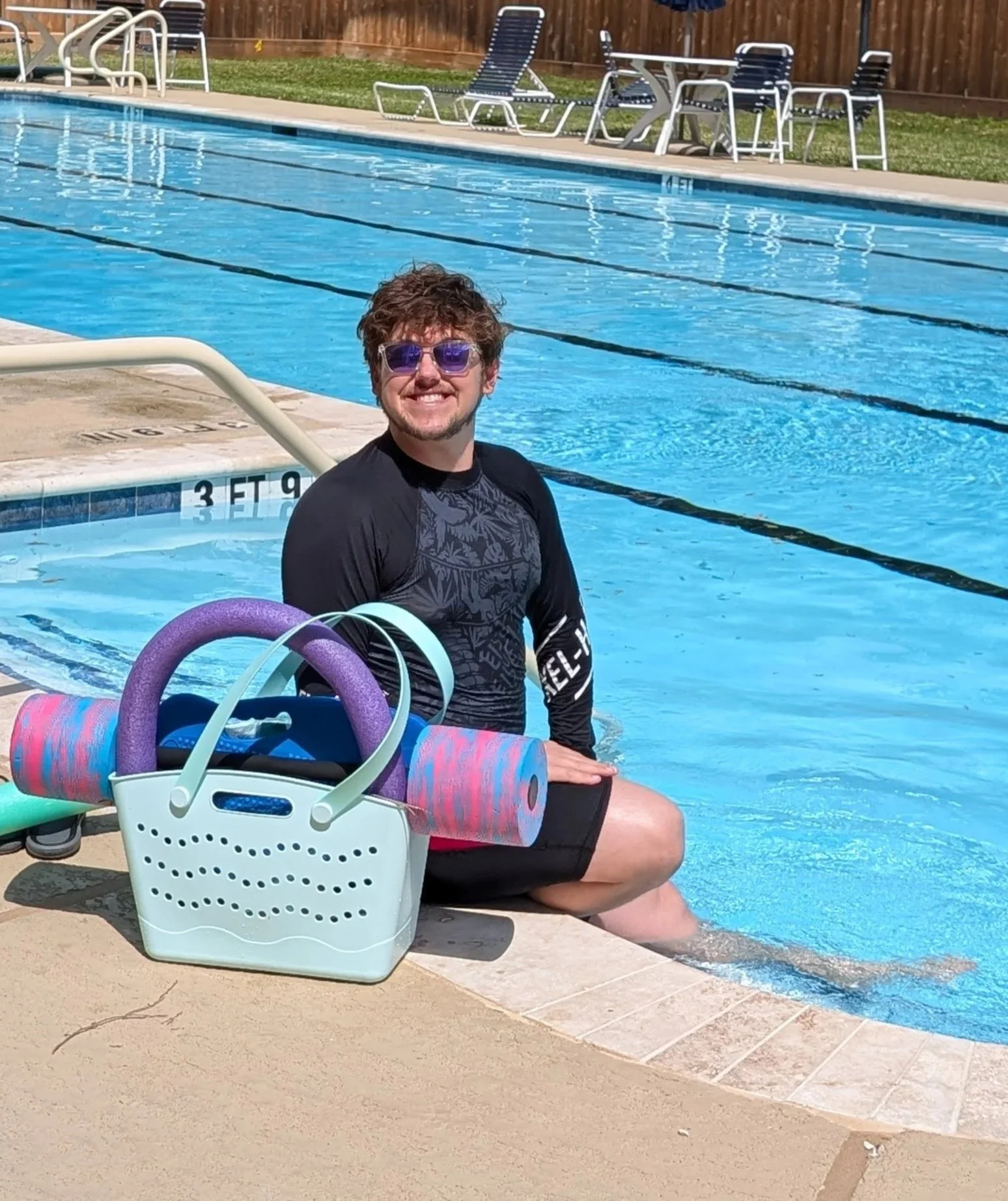 A man sitting on the edge of a swimming pool, smiling, wearing sunglasses and a black rash guard. In front of him on the pool deck, there is a white basket filled with colorful pool noodles and accessories. The background shows patio furniture and a wooden fence.