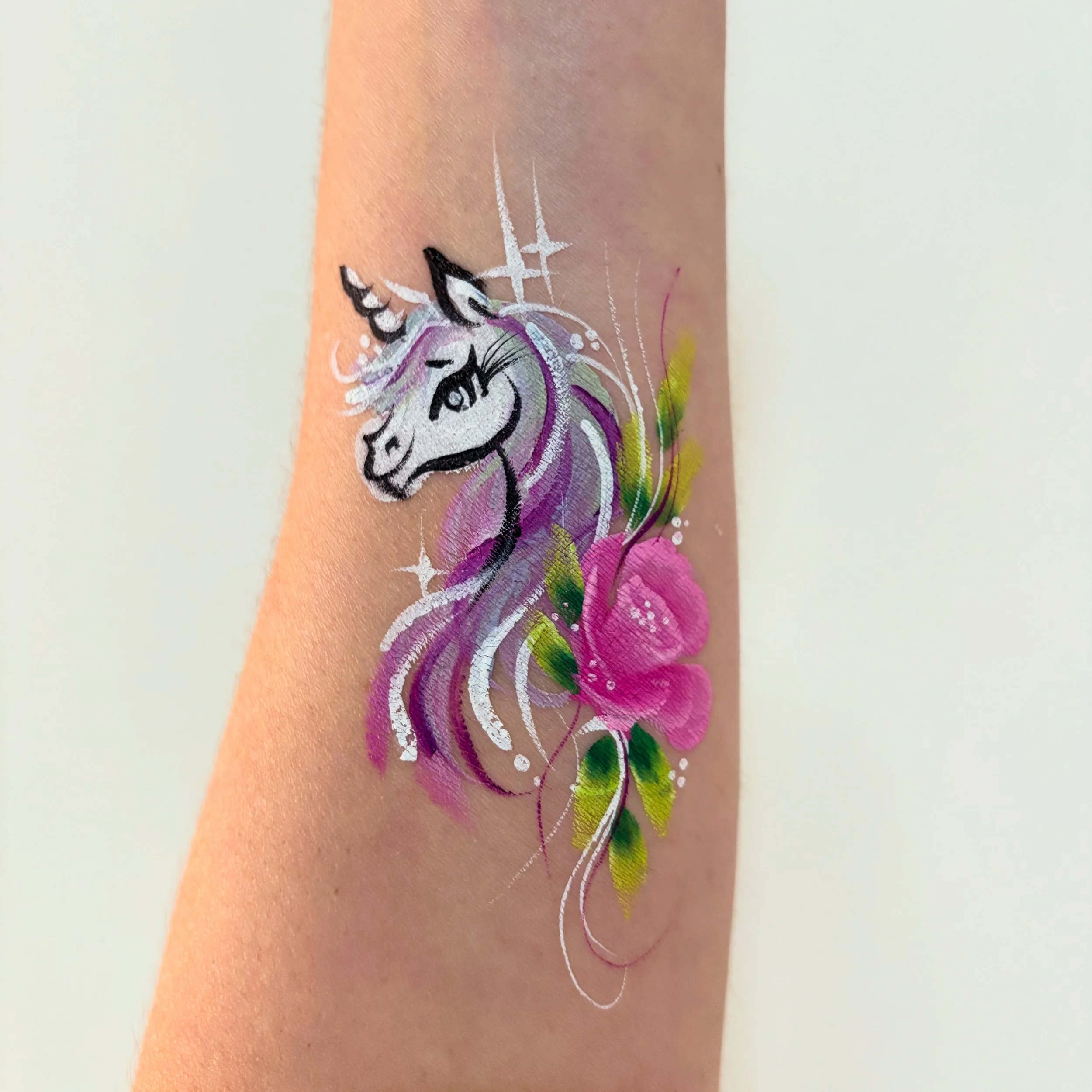 Temporary tattoo of a smiling unicorn head with a pink flower and green leaves on a person's forearm.