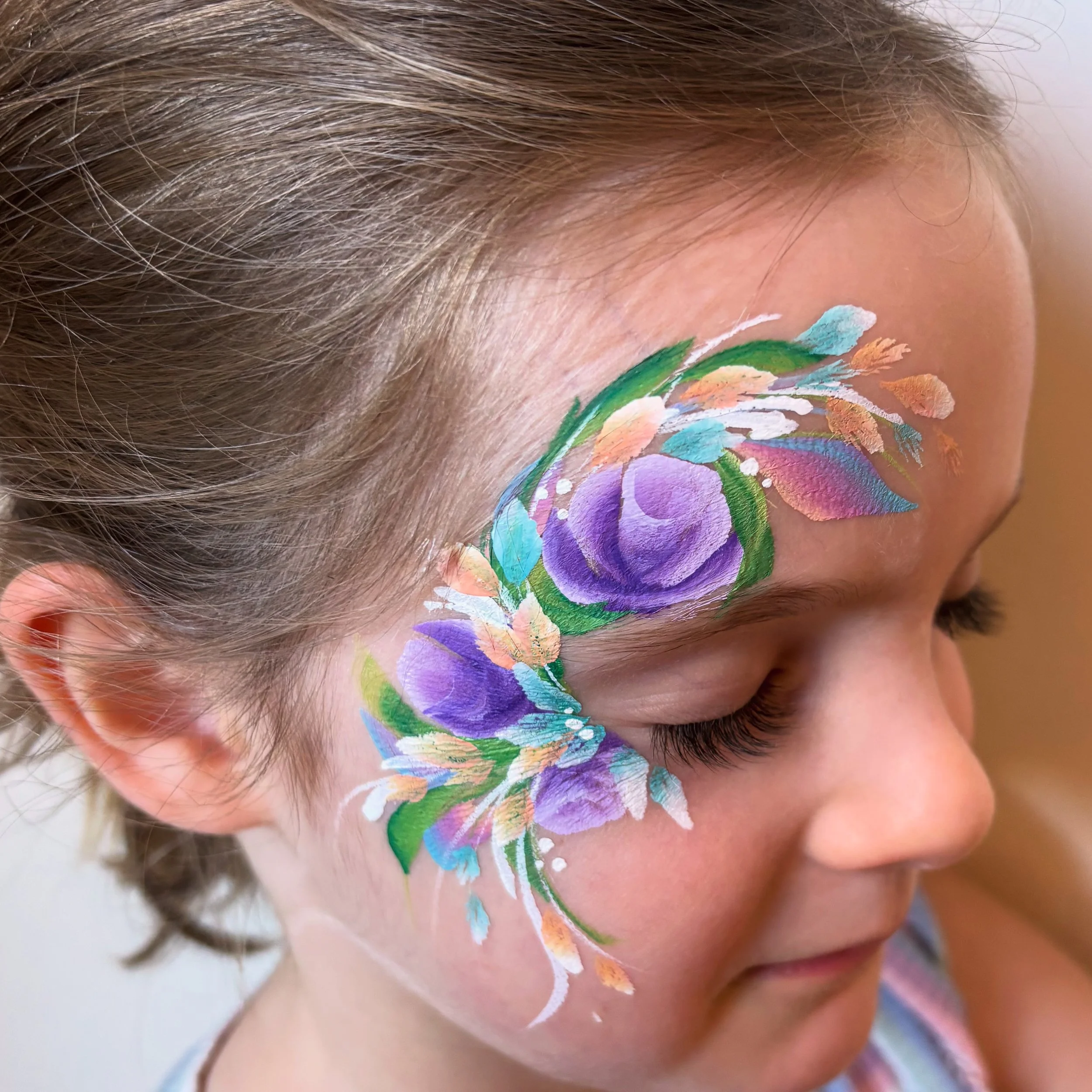 Close-up of a young girl with colorful floral face paint on her forehead and around her eye, featuring purple roses, green leaves, and pastel accents.