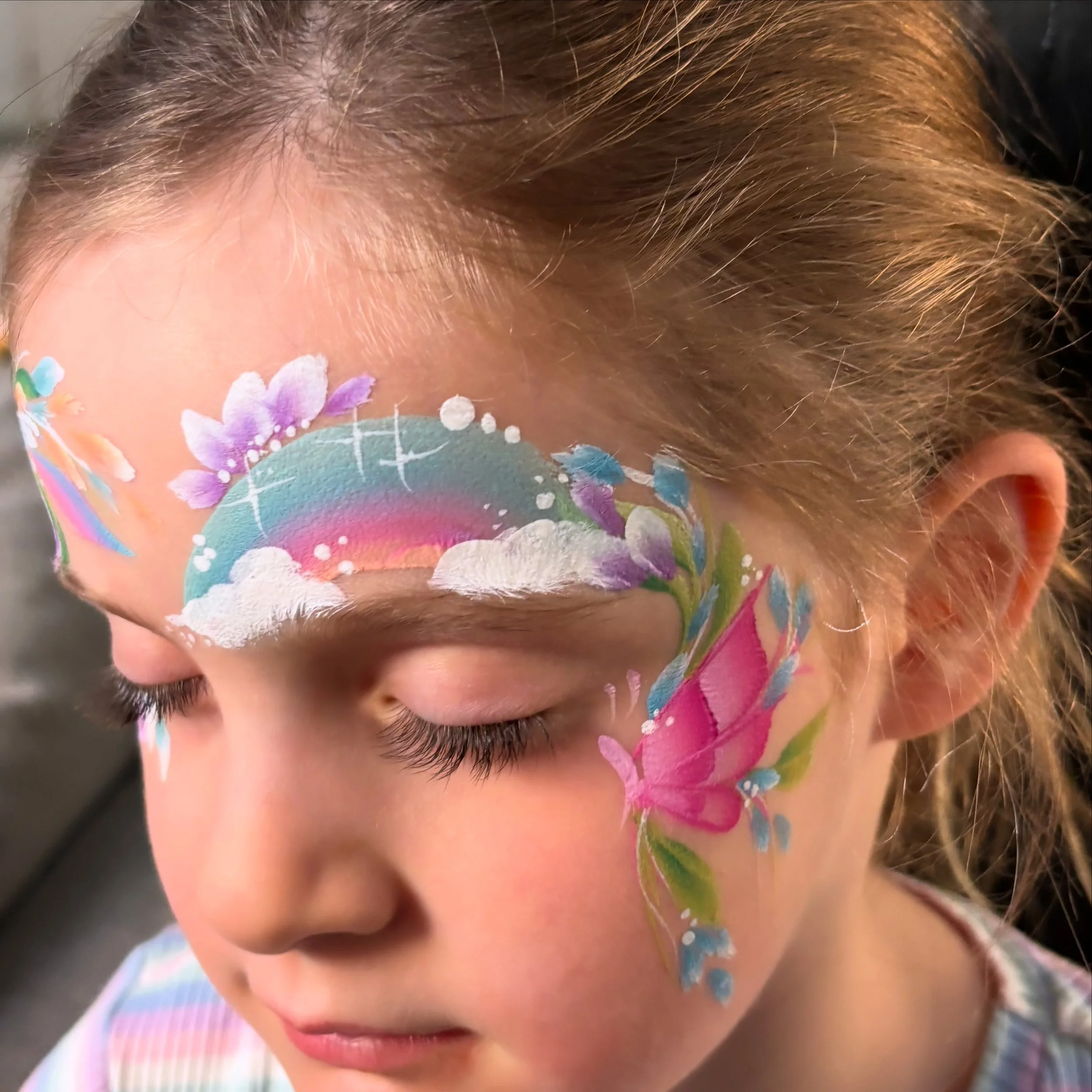 A young girl with face paint of a rainbow, clouds, flowers, a butterfly, and a starry sky on her face.