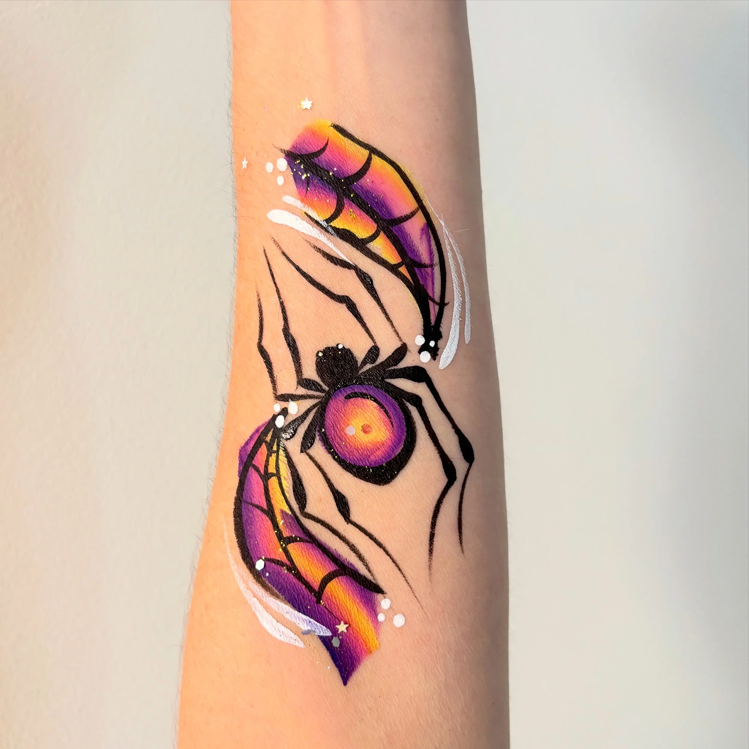 Tattoo of a black widow spider with colorful orange, pink, purple, and yellow accents on its body and two main legs, with spider webs and small white decorative elements surrounding it.
