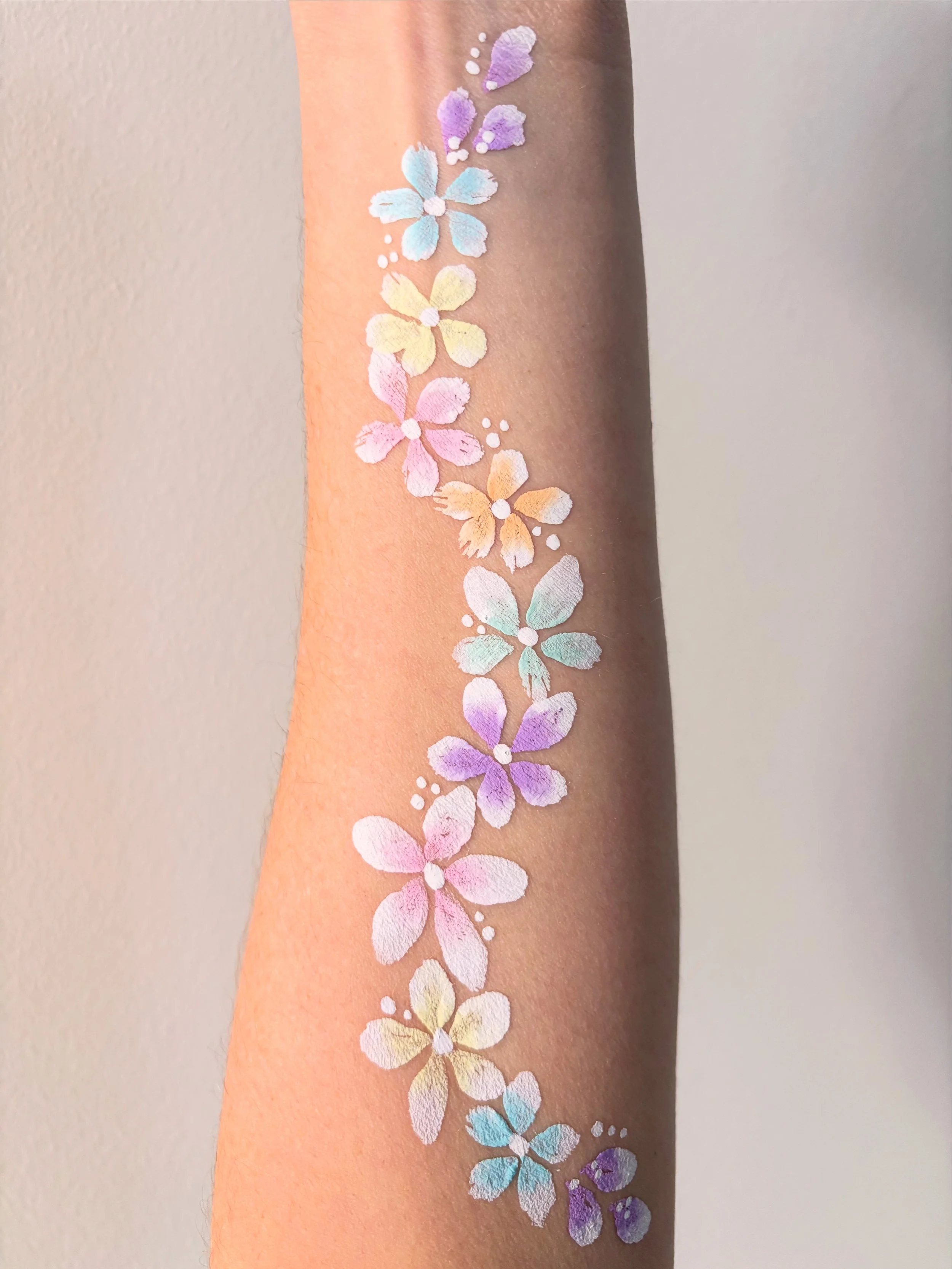 Colorful floral body paint tattoo on forearm featuring pastel-colored flowers in shades of purple, pink, yellow, blue, and white with decorative dots.