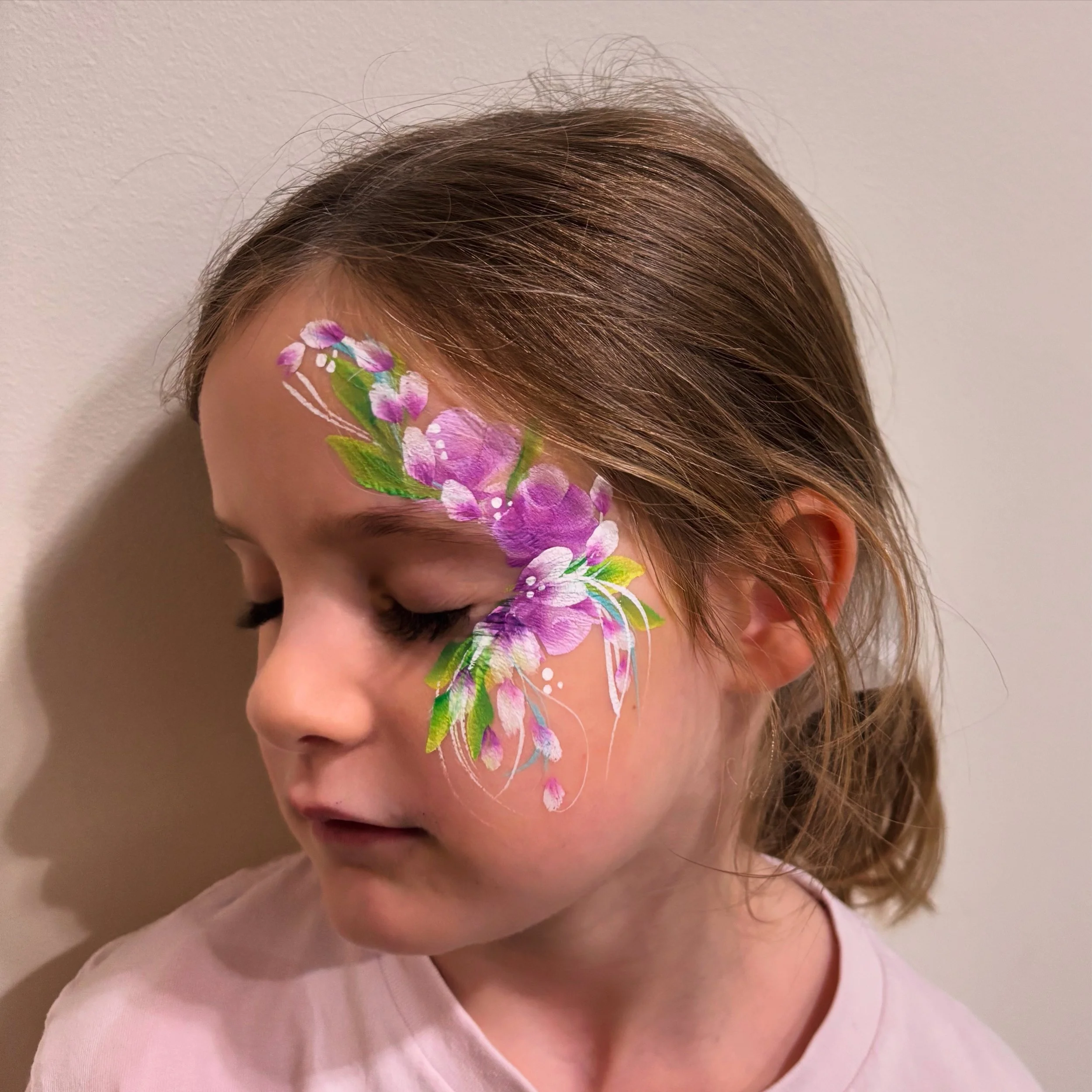 Child with painted purple and white flower design on the right side of her face, closed eyes, light brown hair, wearing a light pink shirt, standing against a plain wall.