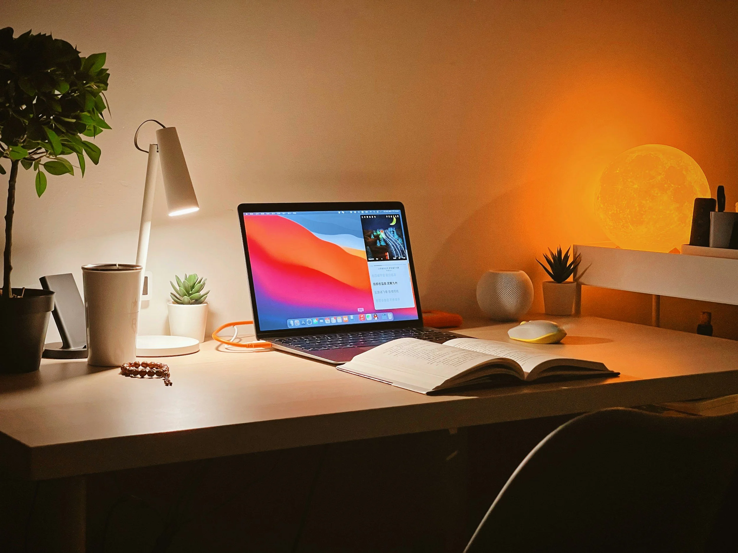 A well-organized desk illuminated by a white lamp, featuring a laptop, open book, and various potted plants with warm lighting, including a LED moon lamp and a moon-themed light casting an orange glow.