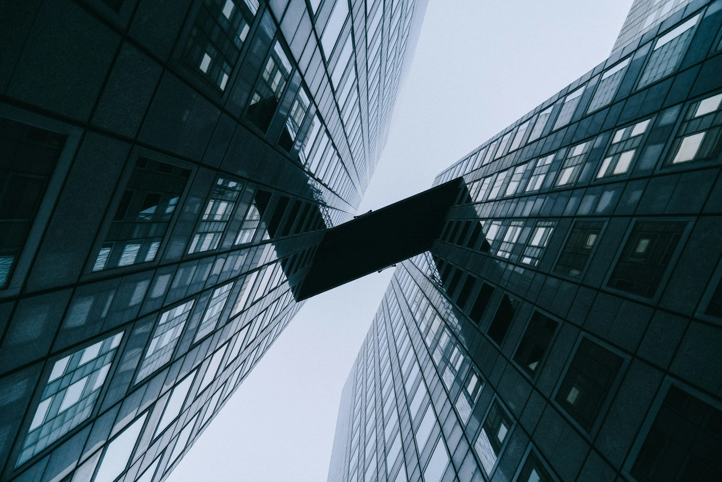 Looking up at two modern glass skyscrapers with a connecting bridge between them, on a cloudy day.