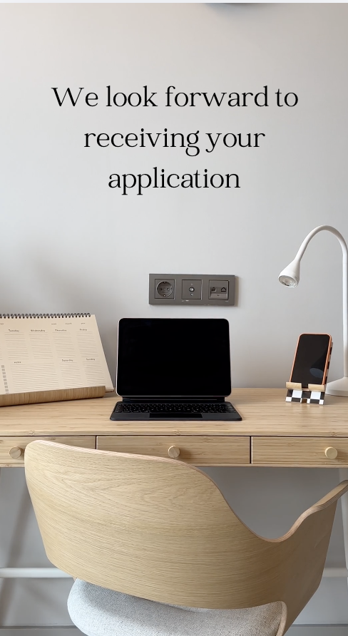 A cozy workspace with a wooden desk, a closed laptop, a standing lamp, a power socket with outlets, a planner, and a smartphone on a stand. There's a quote on the wall saying, "We look forward to receiving your application."