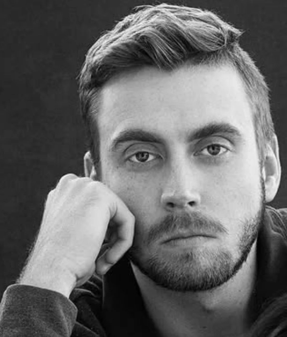 Black and white portrait of a young man with styled hair and a beard, resting his head on his hand and looking directly at the camera.