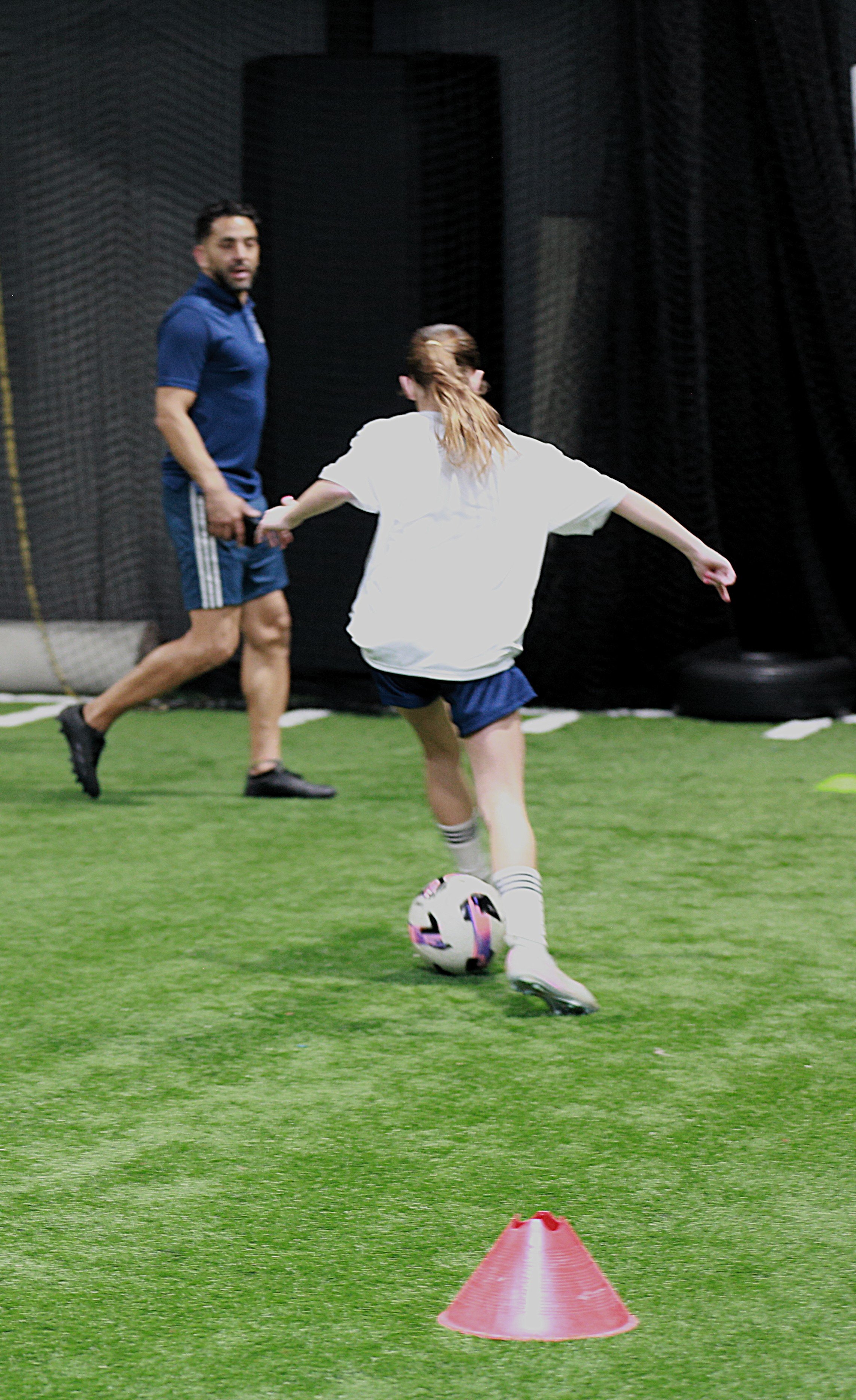 A child in a white shirt and shorts practicing soccer drills on an indoor turf field, dribbling a soccer ball around orange cones under the supervision of an adult coach.