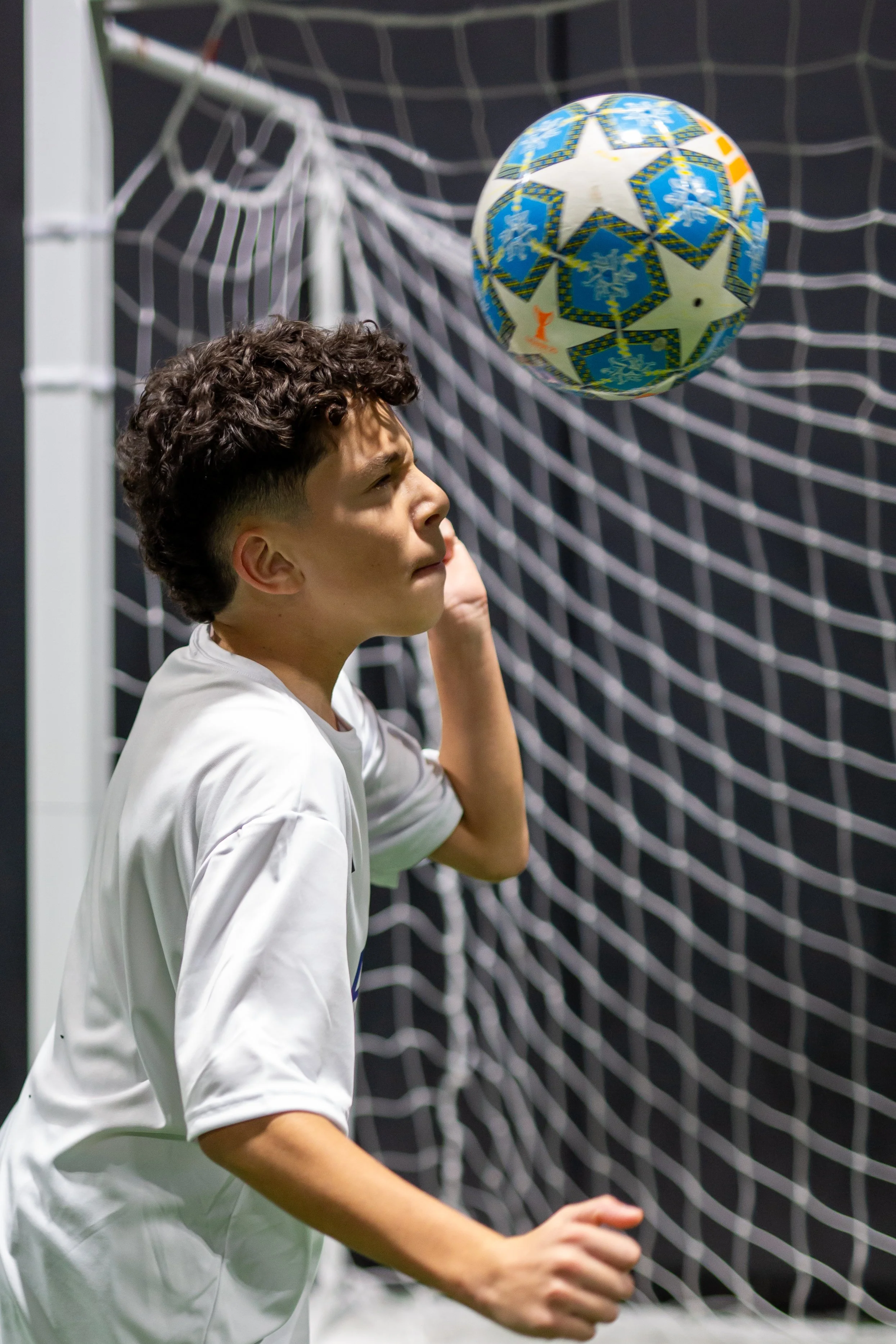 A young boy with curly hair wearing a white sports jersey playfully heads a blue and white soccer ball decorated with stars and snowflakes in front of a goal net.