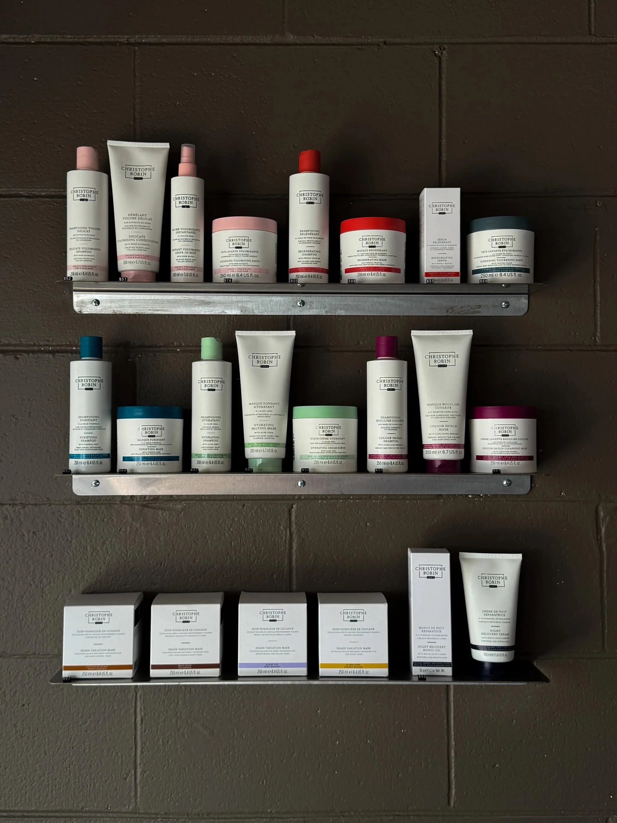 Shelf with various Christophe Robin hair care products, including shampoos, conditioners, masks, and treatments, arranged on three levels against a brown wall.