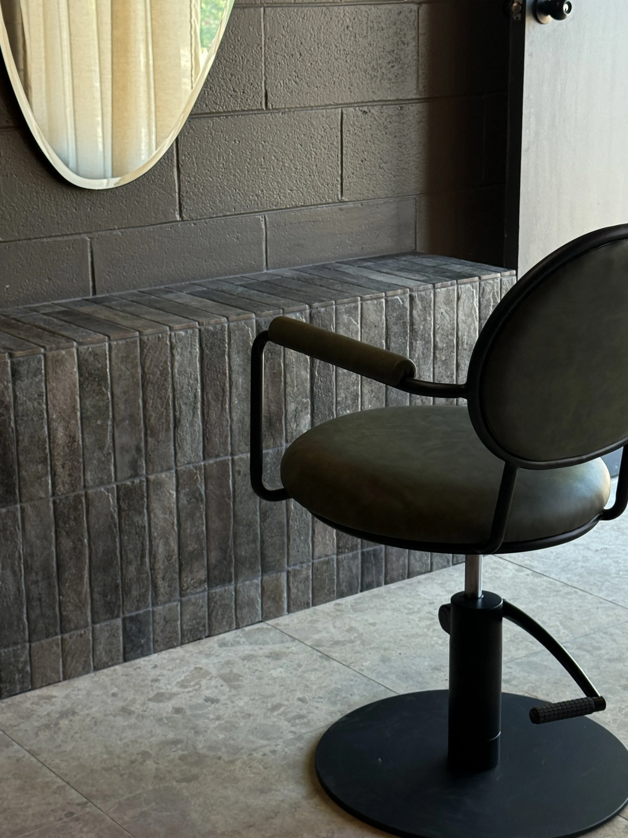 An empty salon or barber chair with black frame and green seat, positioned in front of a mirror on a textured grey wall with tiled ledge, on a tiled floor.