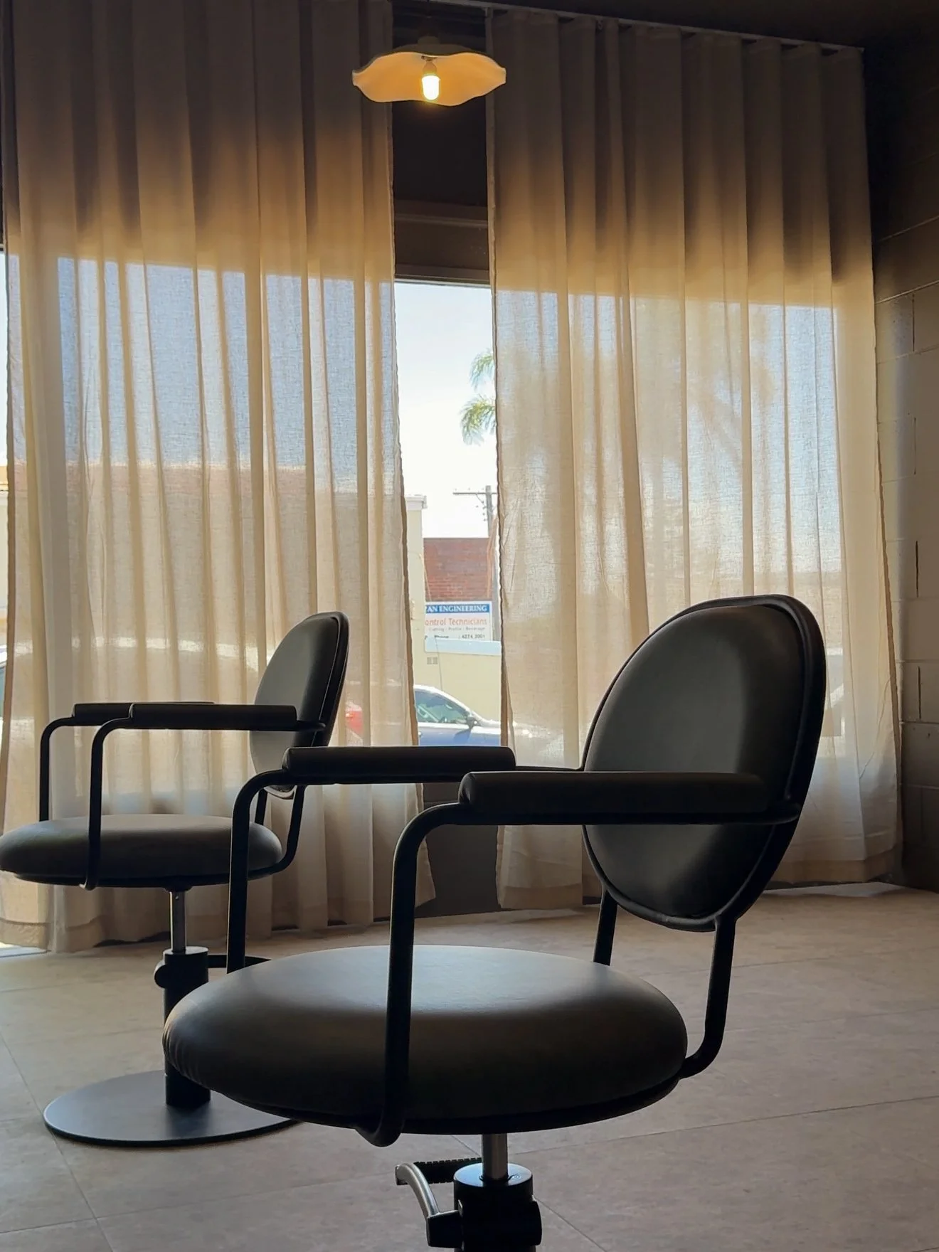 Interior scene with two black office chairs facing away from large window with beige curtains, ceiling light, and partly visible outside view of a car and a building.