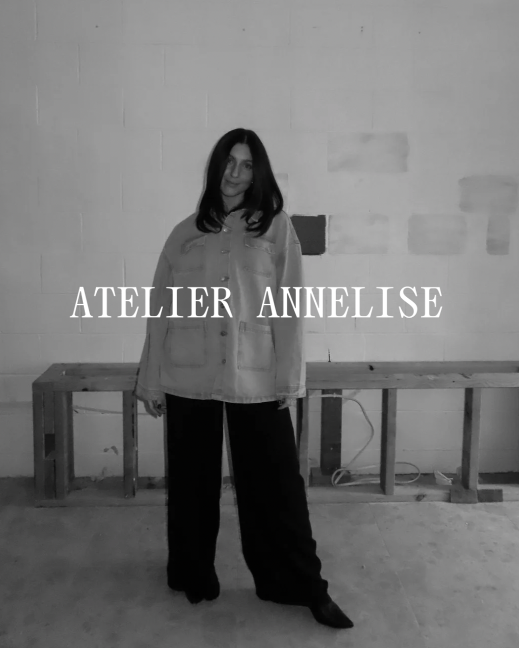 Black and white photo of a woman standing in front of a plain wall, wearing a jacket and wide-leg pants, with the text 'ATELIER ANNELISE' across the center.