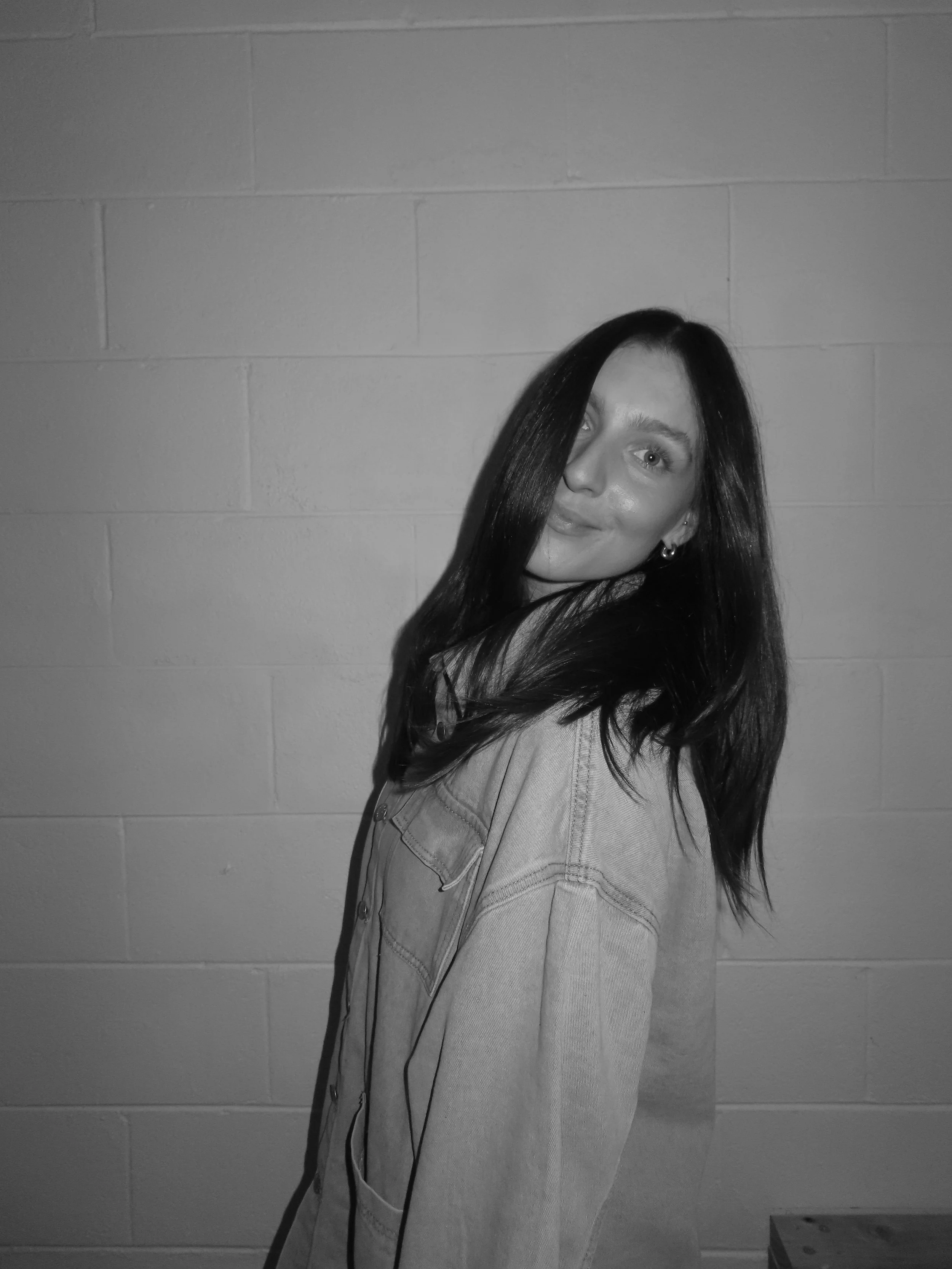 A woman with long dark hair smiling, standing against a plain wall, wearing a casual light-colored denim jacket.
