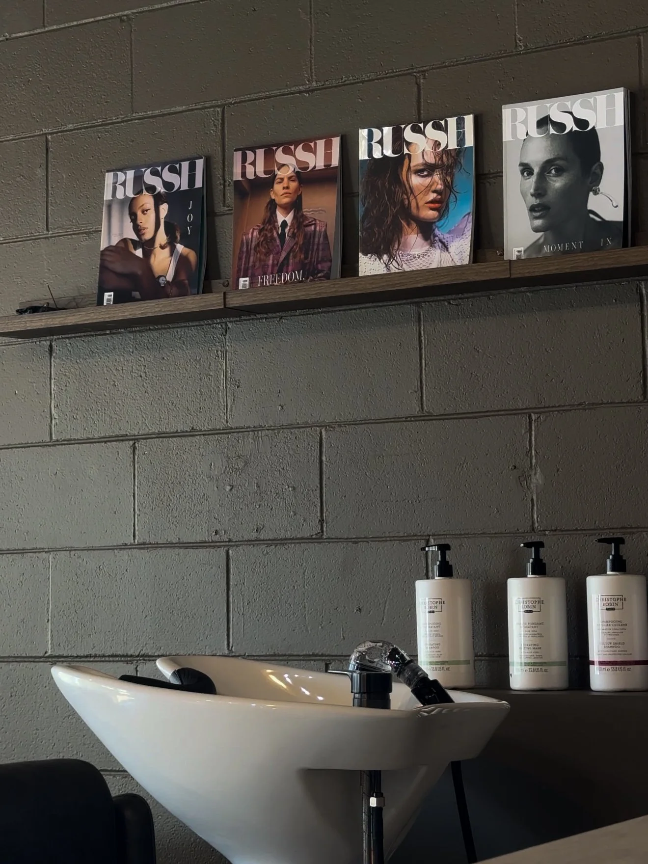 Display of four fashion magazines on a wooden shelf above a hair-washing station with a white sink and bottles of hair products on the side.