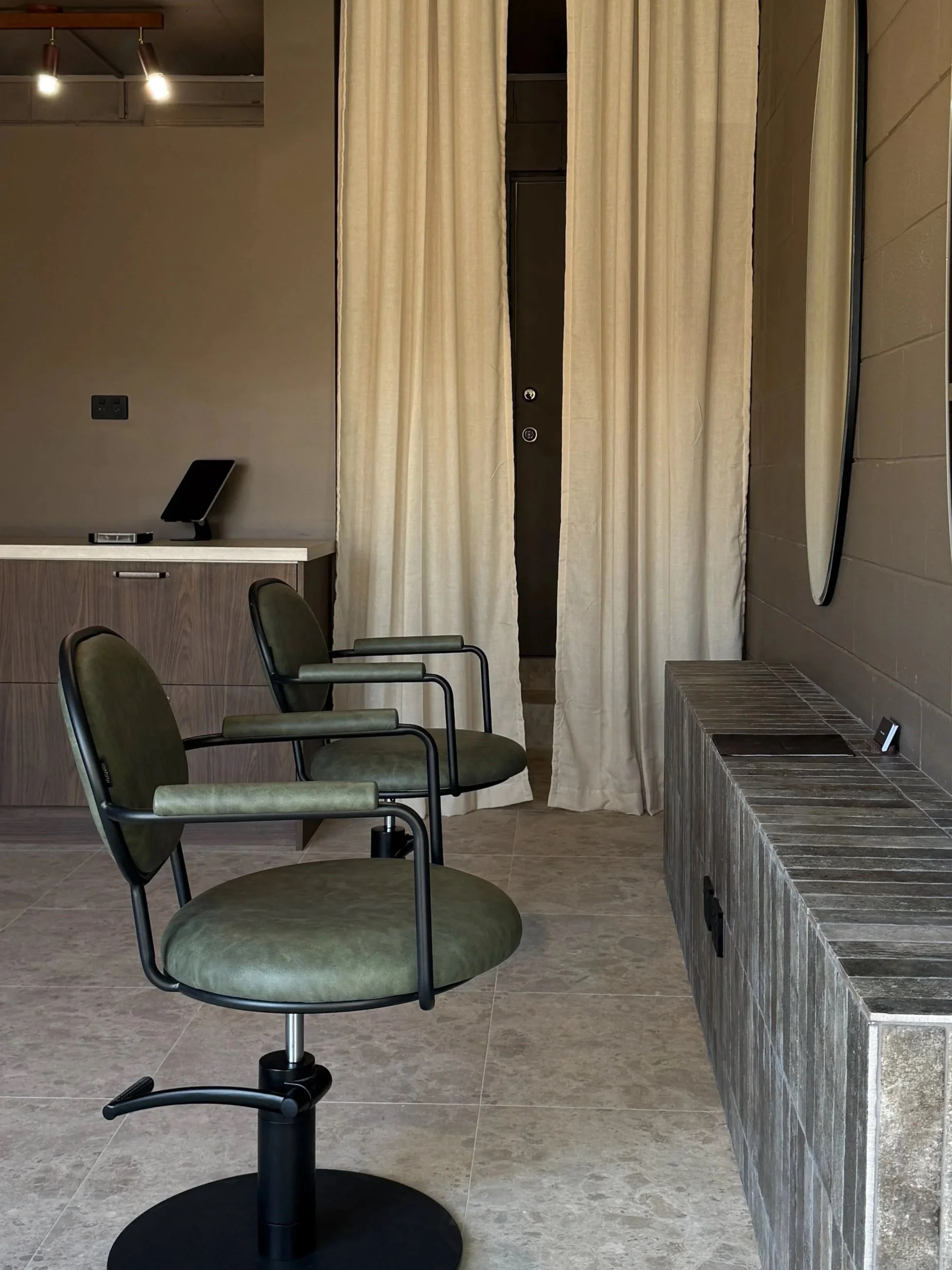 Interior of a modern room with two green office chairs on wheels, a wooden cabinet, a curtain covering a door or window, a wall-mounted mirror, and tiled flooring.