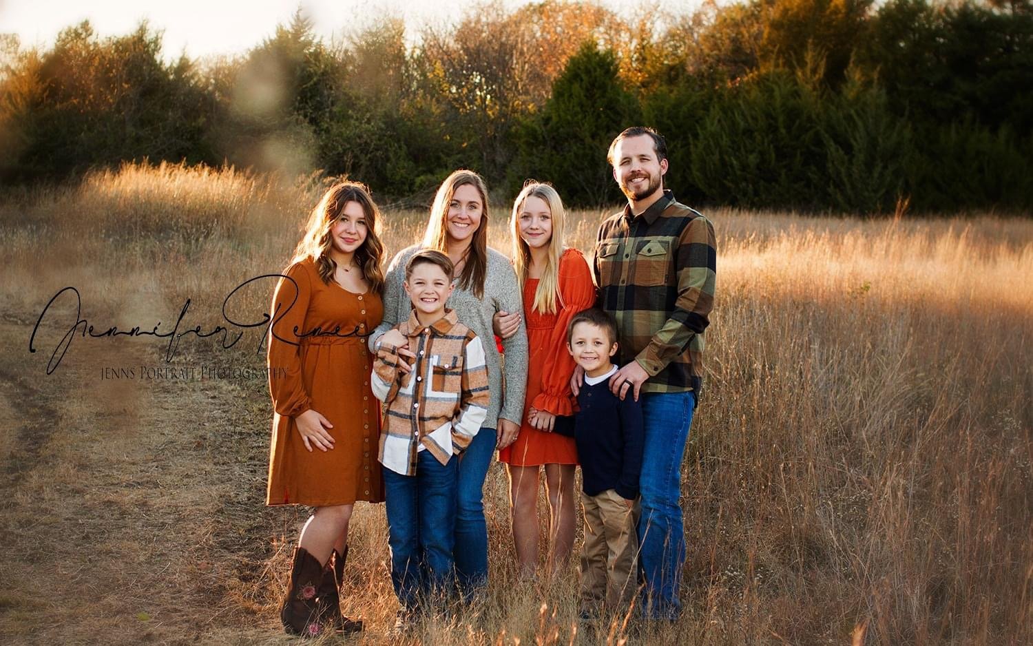 Family of six standing on a dirt path in a field during sunset, smiling and dressed in fall clothing, with trees in the background.