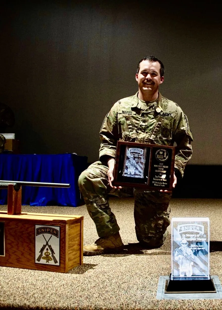 A man in military uniform kneeling on one knee holding a plaque, smiling at the camera during an award ceremony, with a wooden box and a trophy with sniper insignia nearby.