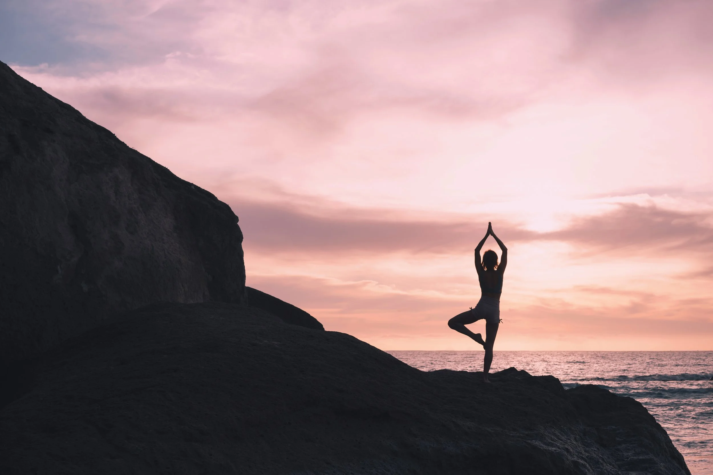 A person practicing yoga in tree pose on a rocky terrain during sunset by the ocean, with pink and purple sky and clouds.