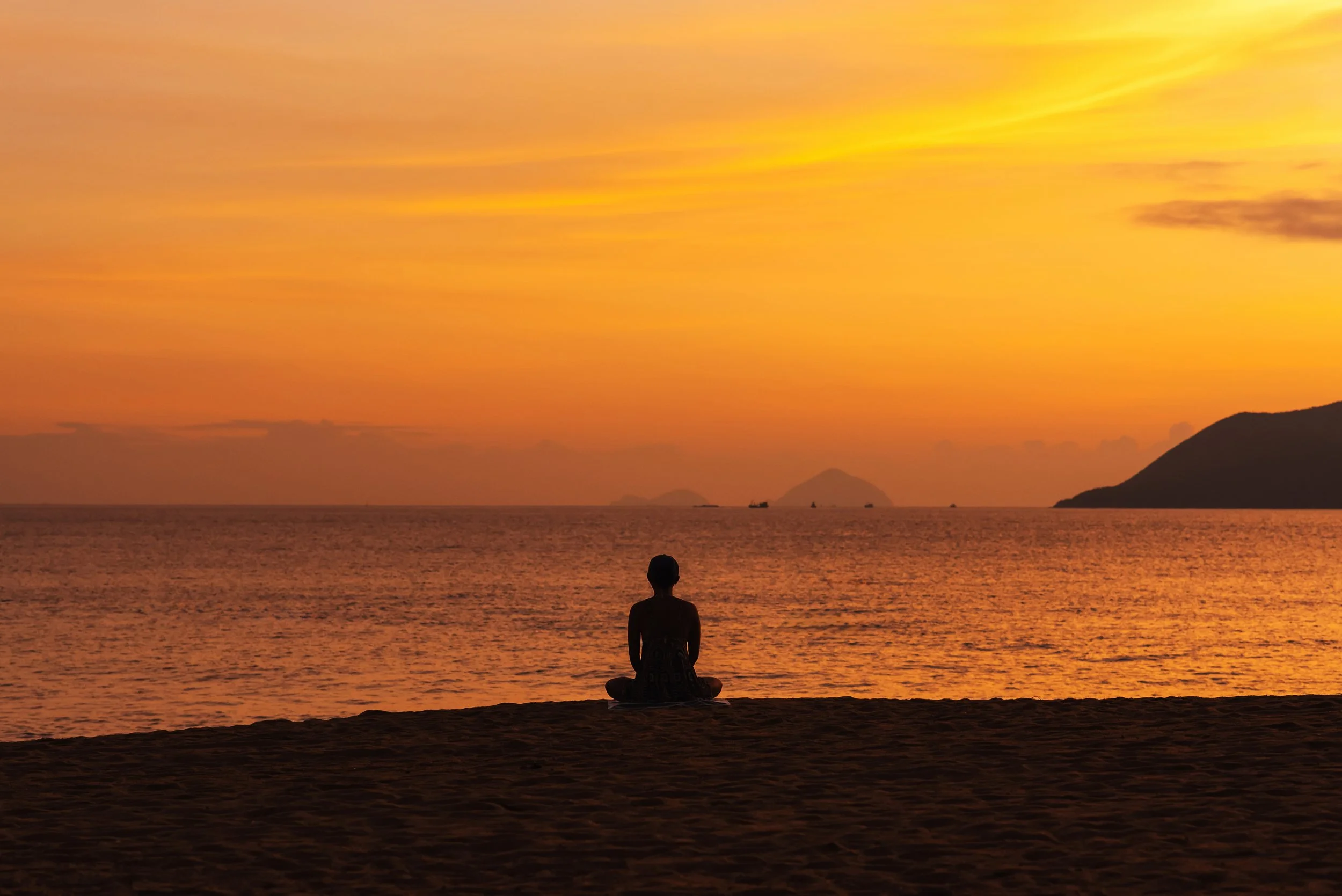 A person sitting cross-legged on the beach watching a vibrant sunset over the ocean with distant islands and mountains.