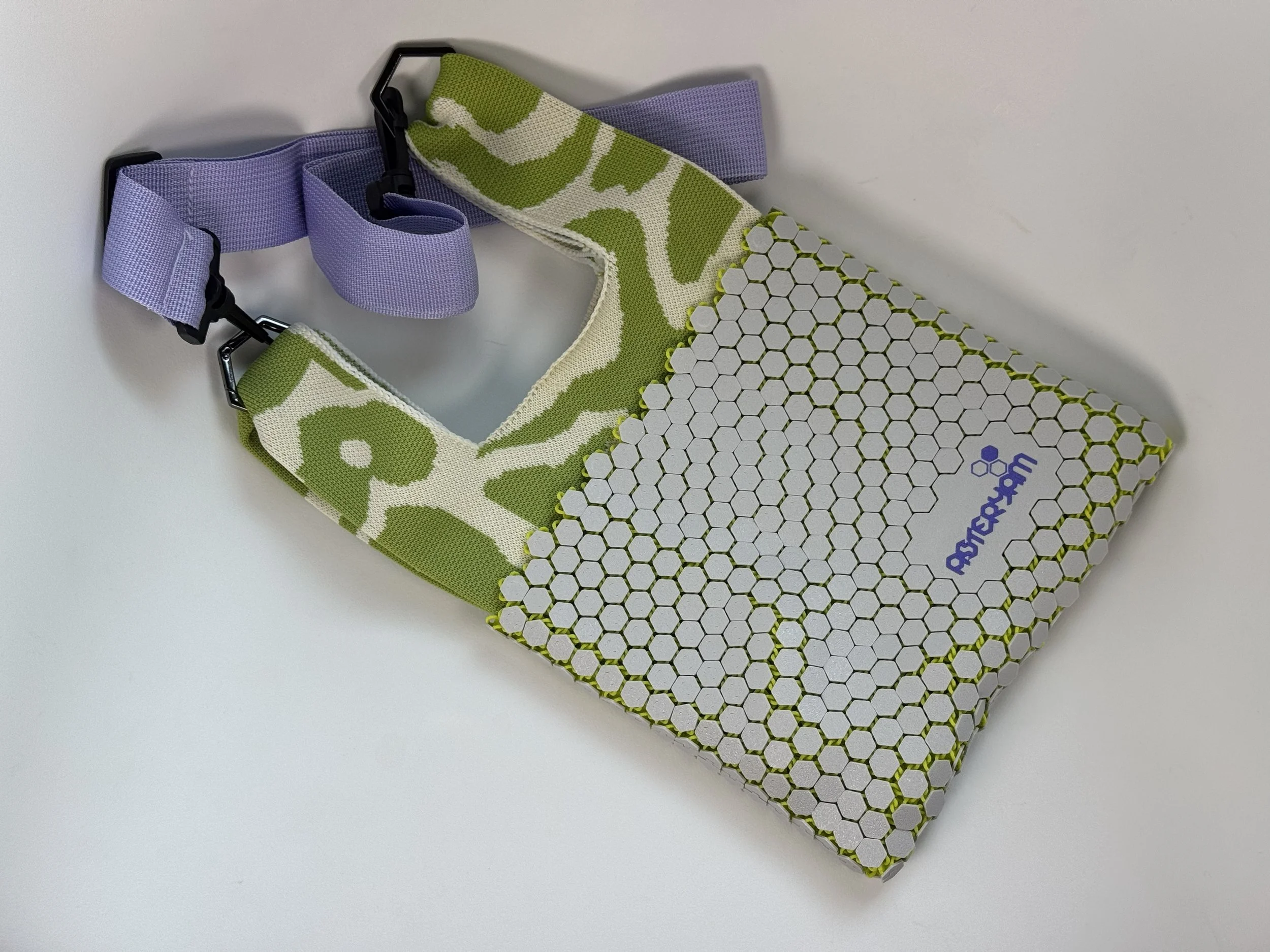 Flat lay of ASTERYAM Hexa Sling 3D printed bag. Grey hexagonal mesh shell with green and white knitted lining and purple adjustable crossbody strap.