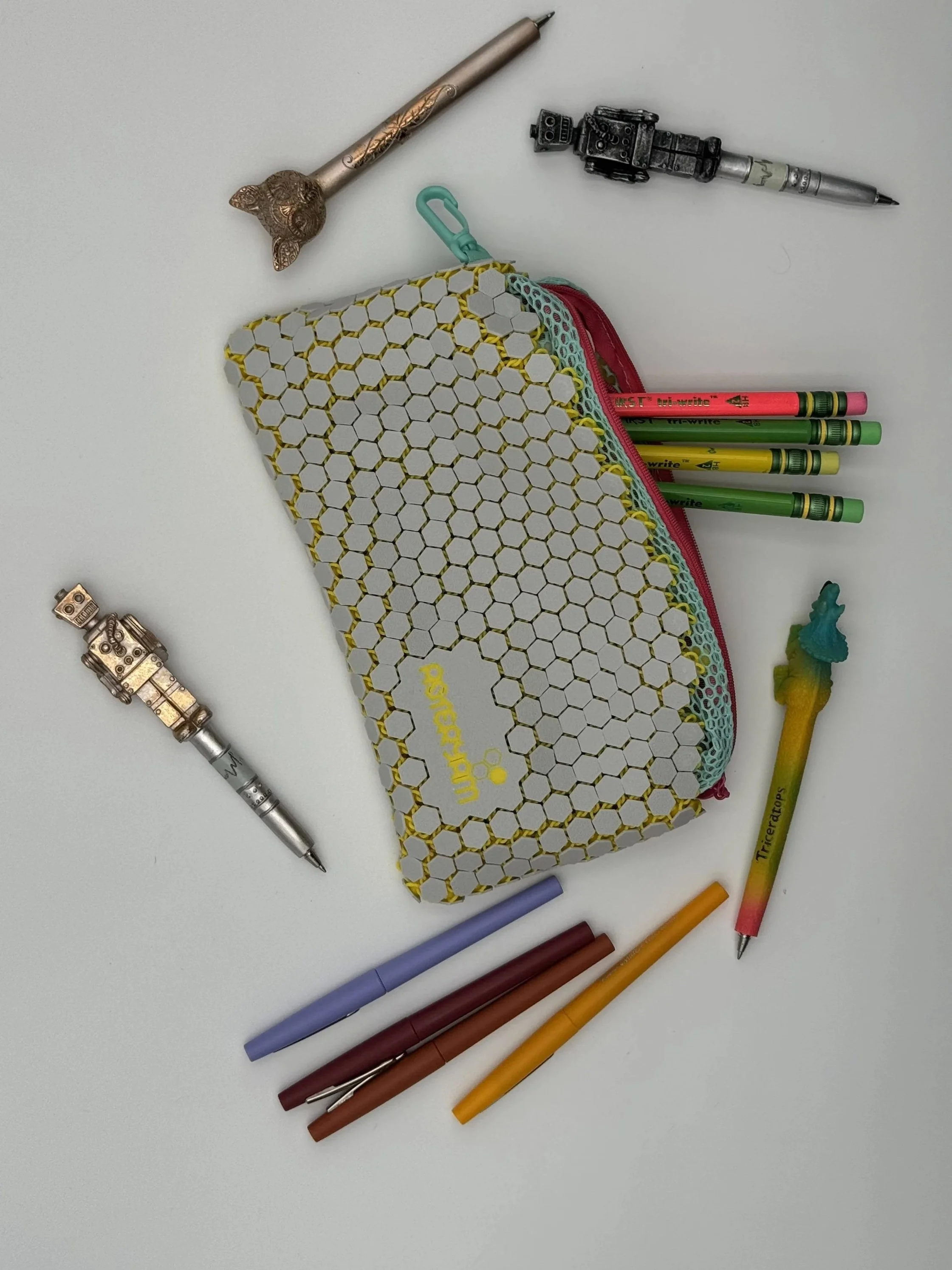 Flat lay of ASTERYAM HexaFlux hexagonal mesh bag used as a pencil case. The grey and yellow bag is surrounded by pens, markers, and vintage robot figurines.
