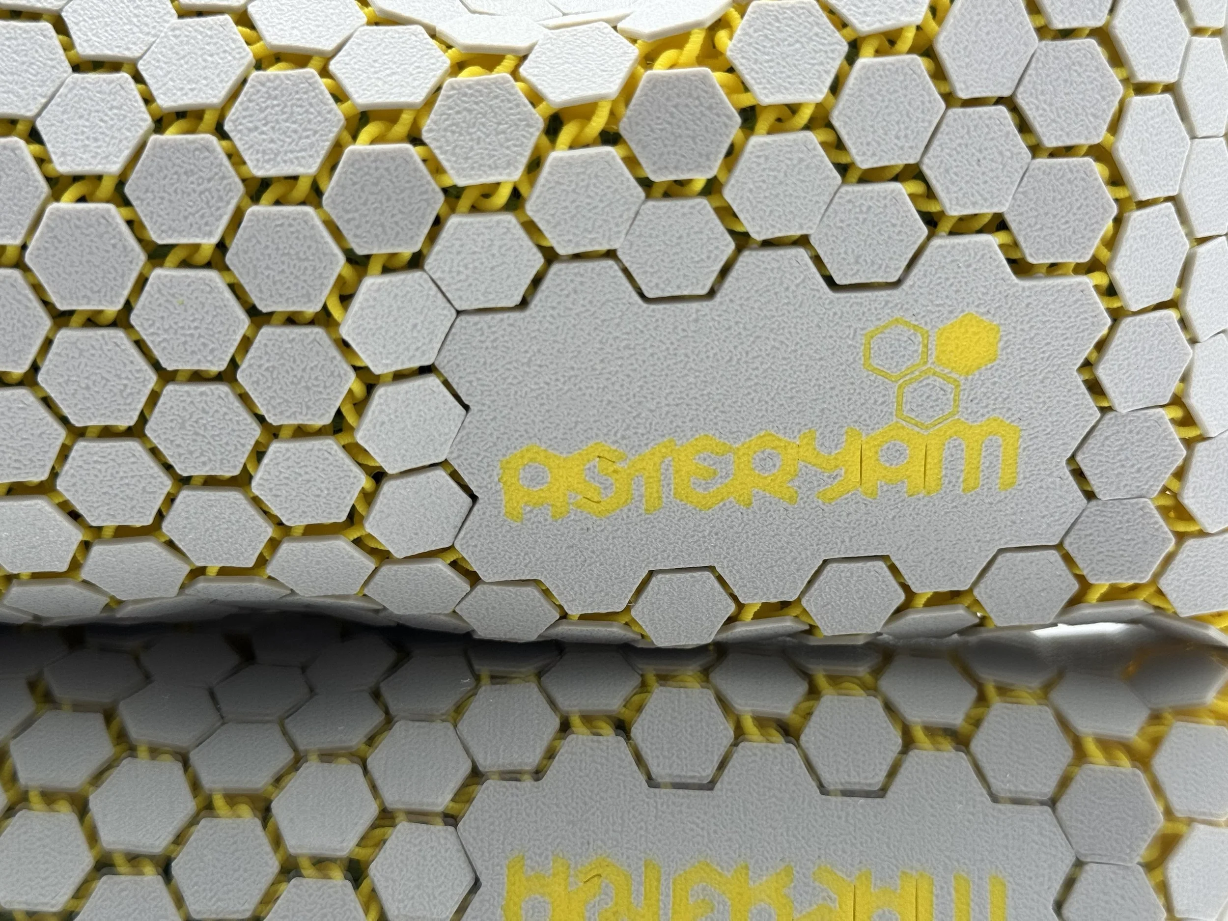 Close-up flat lay of the ASTERYAM HexaFlux bag showing the yellow embossed logo and the precision of the interlocking light grey hexagonal tiles.