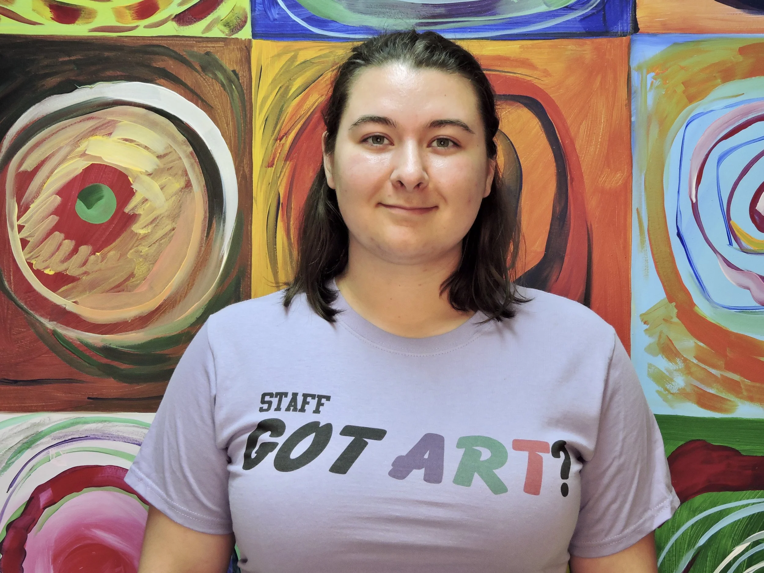 A young woman with shoulder-length dark hair and light skin standing in front of a colorful abstract mural. She is wearing a light purple T-shirt with the words "Staff Got Art?" in black, purple, green, orange, and red lettering.
