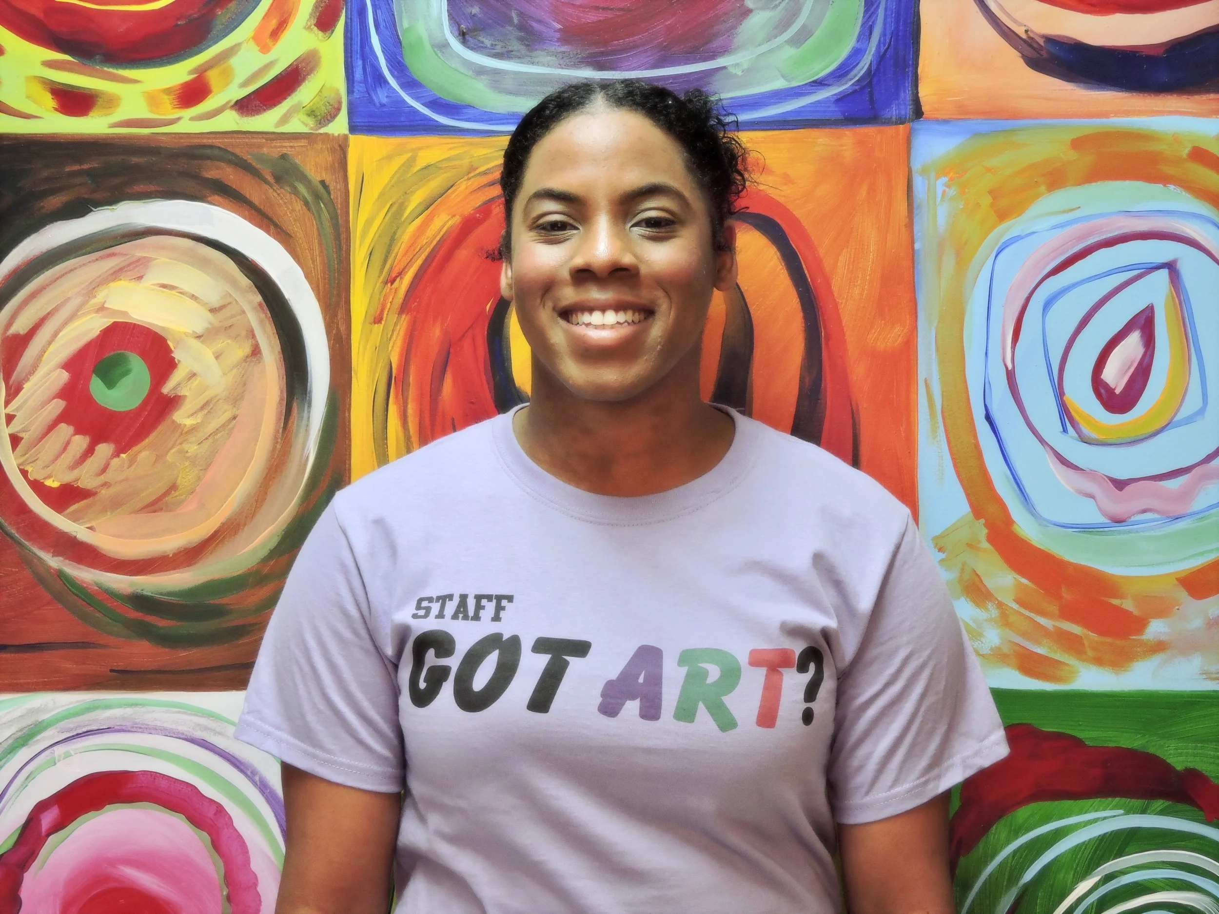 Young man smiling, wearing a light gray T-shirt with the message 'STAFF GOT ART?' in colorful letters, standing in front of a vibrant, abstract, multicolored painted background.