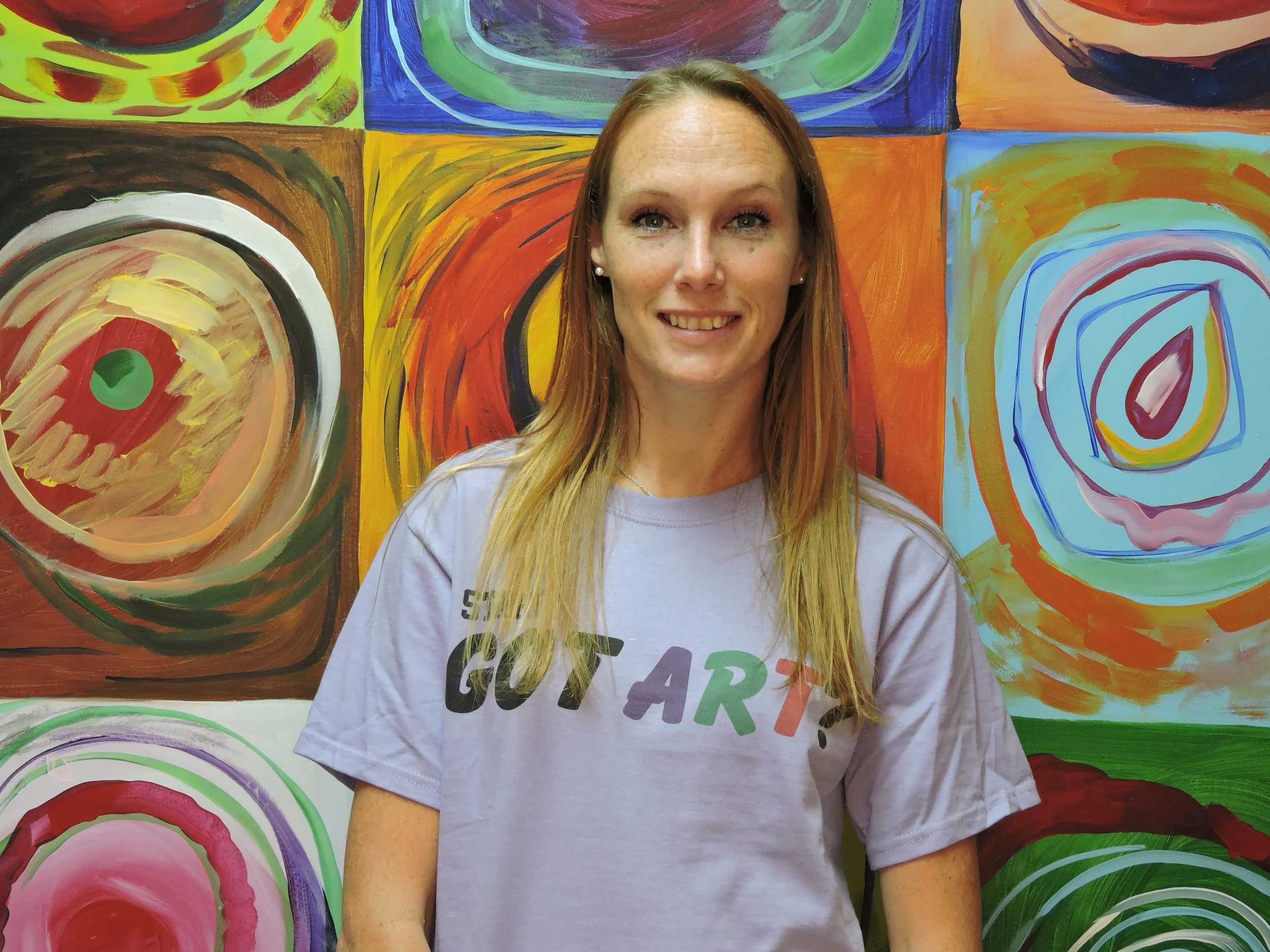 A smiling woman with long blonde hair standing in front of a colorful abstract mural with circular patterns.