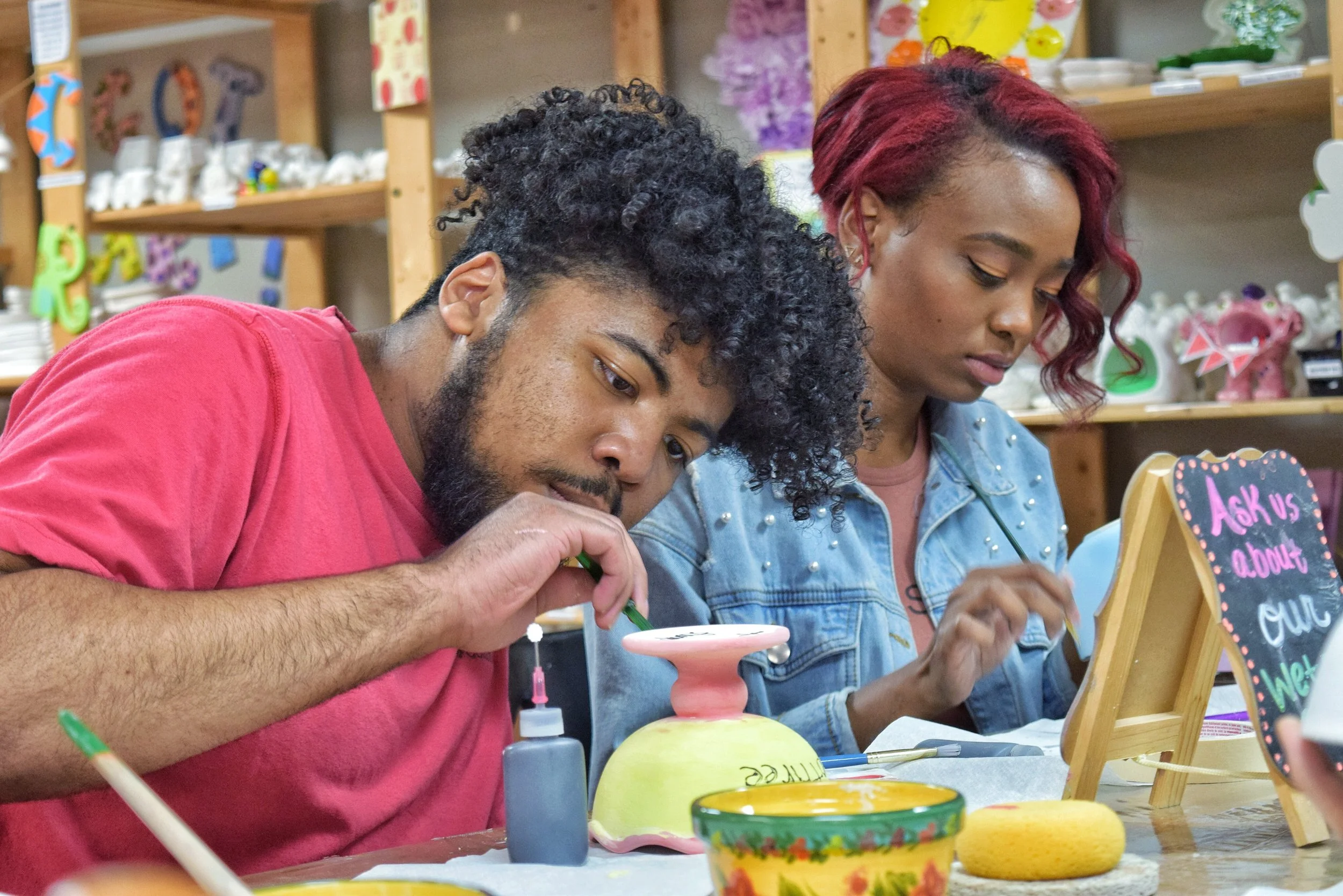 Two people painting ceramic pottery at a craft table, with a sign that says 'Ask us about our!' and shelves with craft supplies in the background.