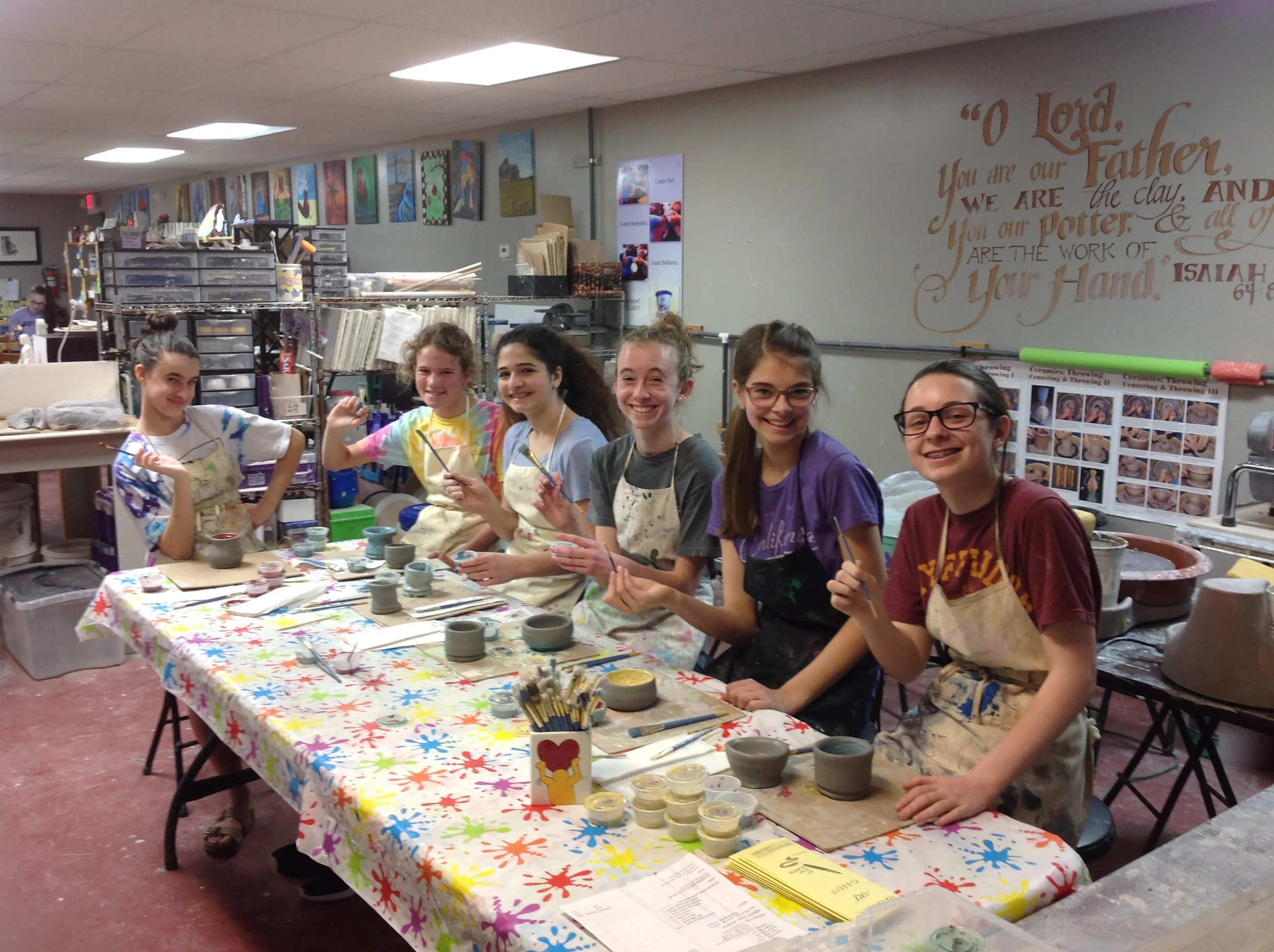 Six young women sit at a table in a pottery studio, smiling and holding brushes with pottery pieces in front of them, surrounded by various pottery supplies and colorful artwork on the walls.