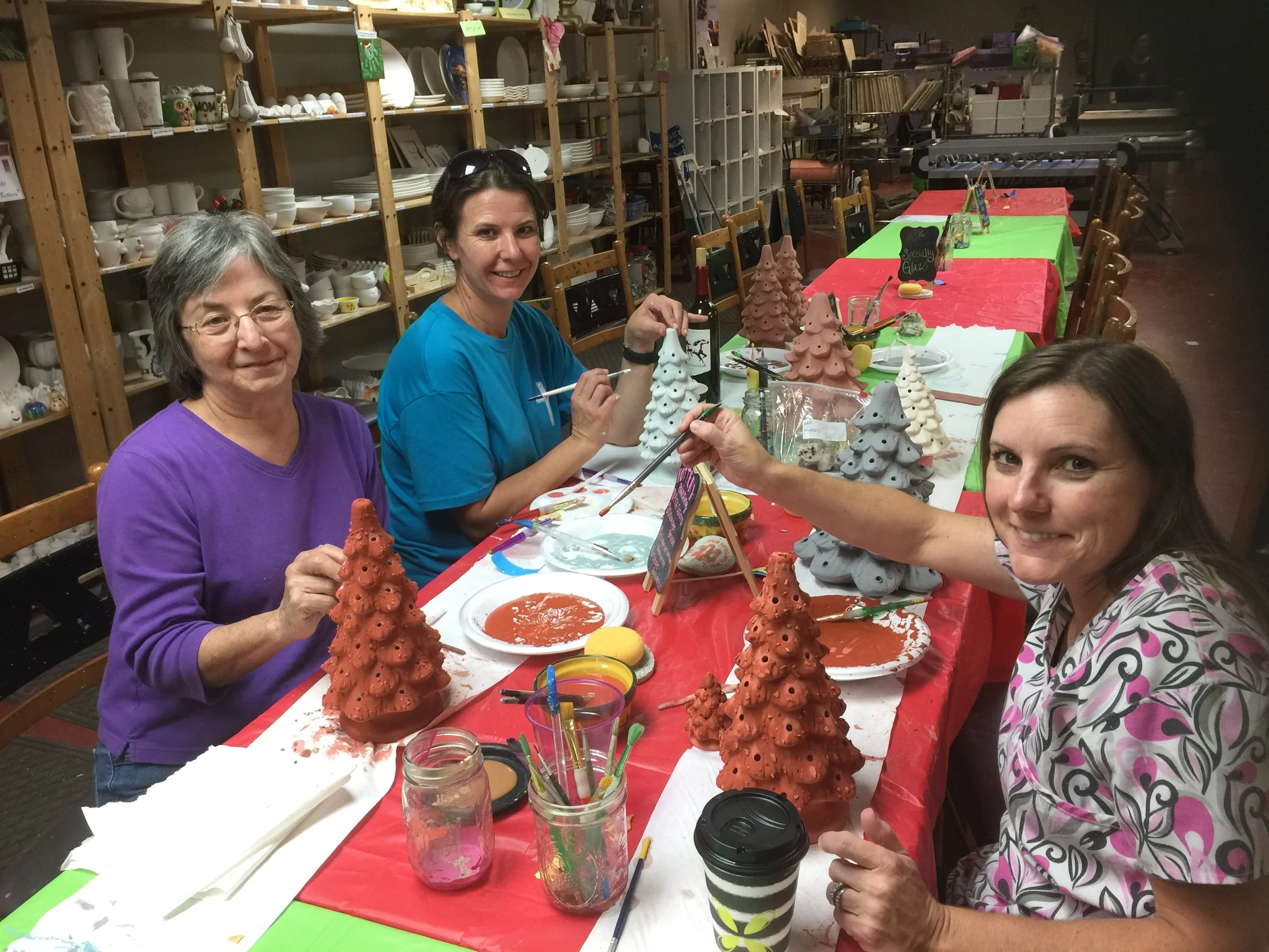 Three women decorating ceramic Christmas trees at a craft table in a ceramics studio.