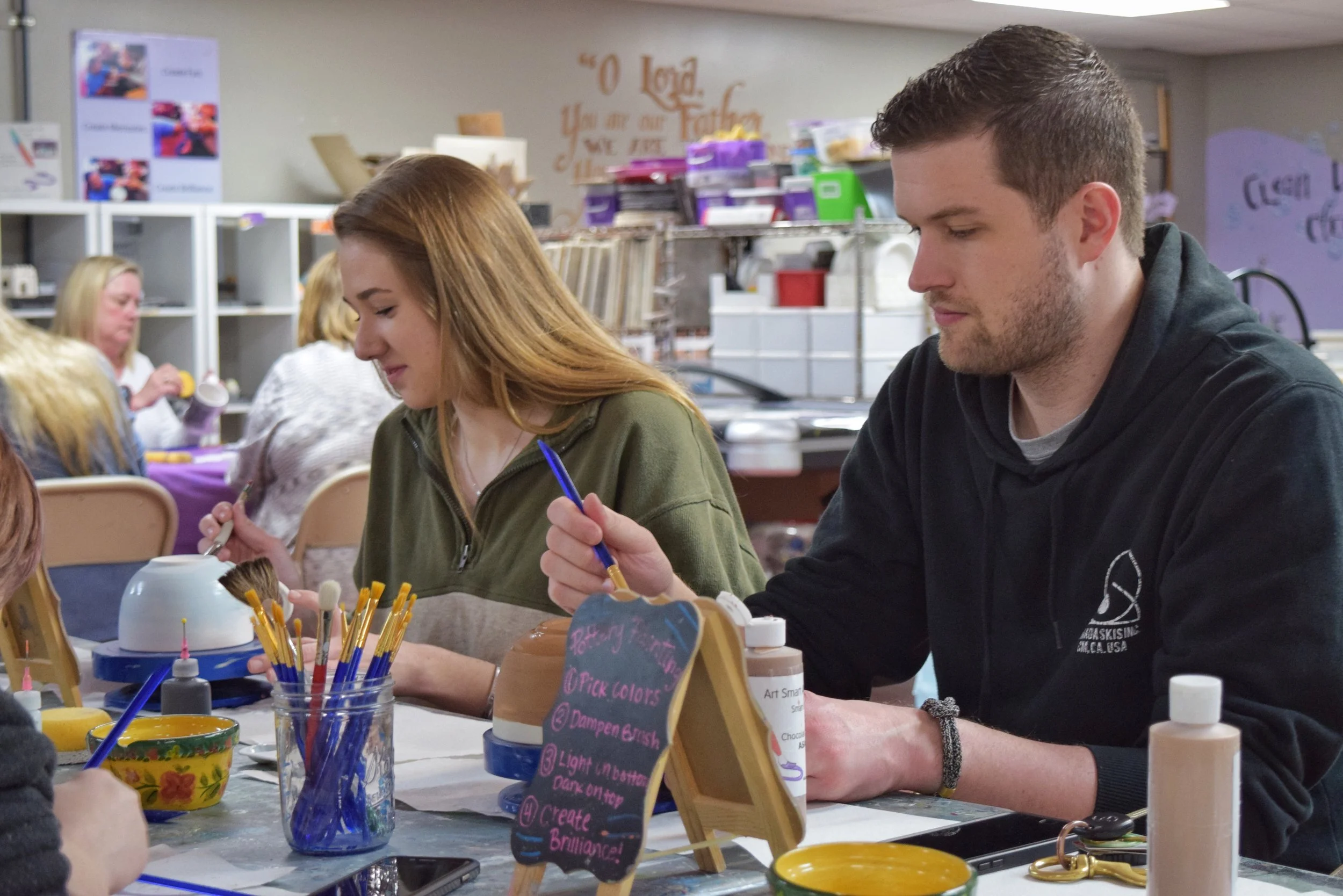 People painting ceramics at a crafting event, with brushes, paint bottles, and ceramic items on the table.