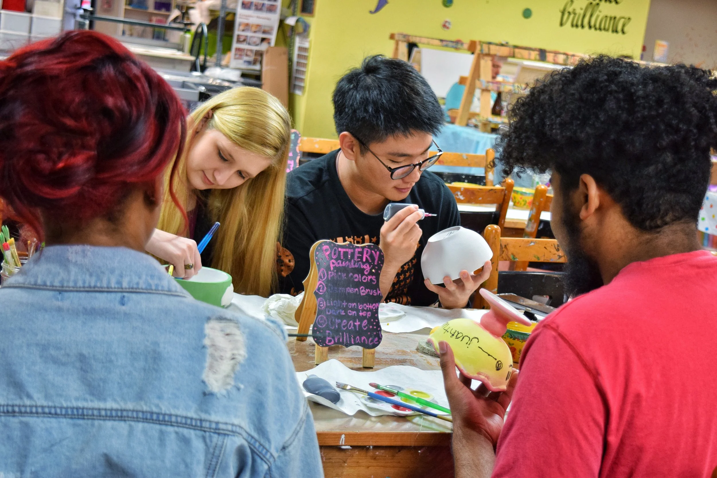 Four individuals are seated at a table, engaged in painting pottery. One person is painting a white ceramic bowl, another is holding a painted ceramic piece with the name 'Jumah' written on it, and two others are focusing on their projects. There is 