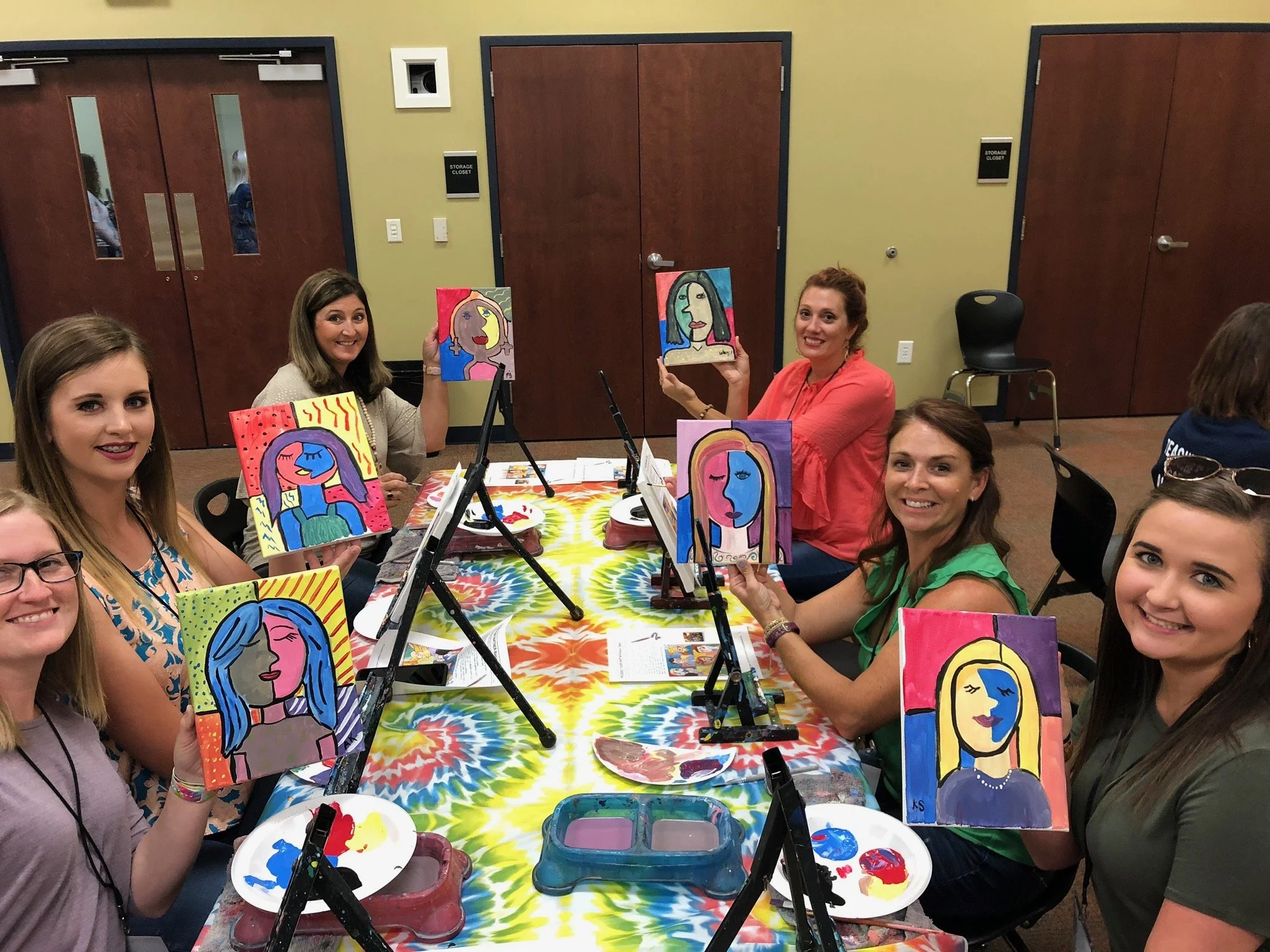 Group of women at a painting party holding colorful portrait paintings they created, sitting at a table with painting supplies, in a room with brown doors and a tie-dye tablecloth.
