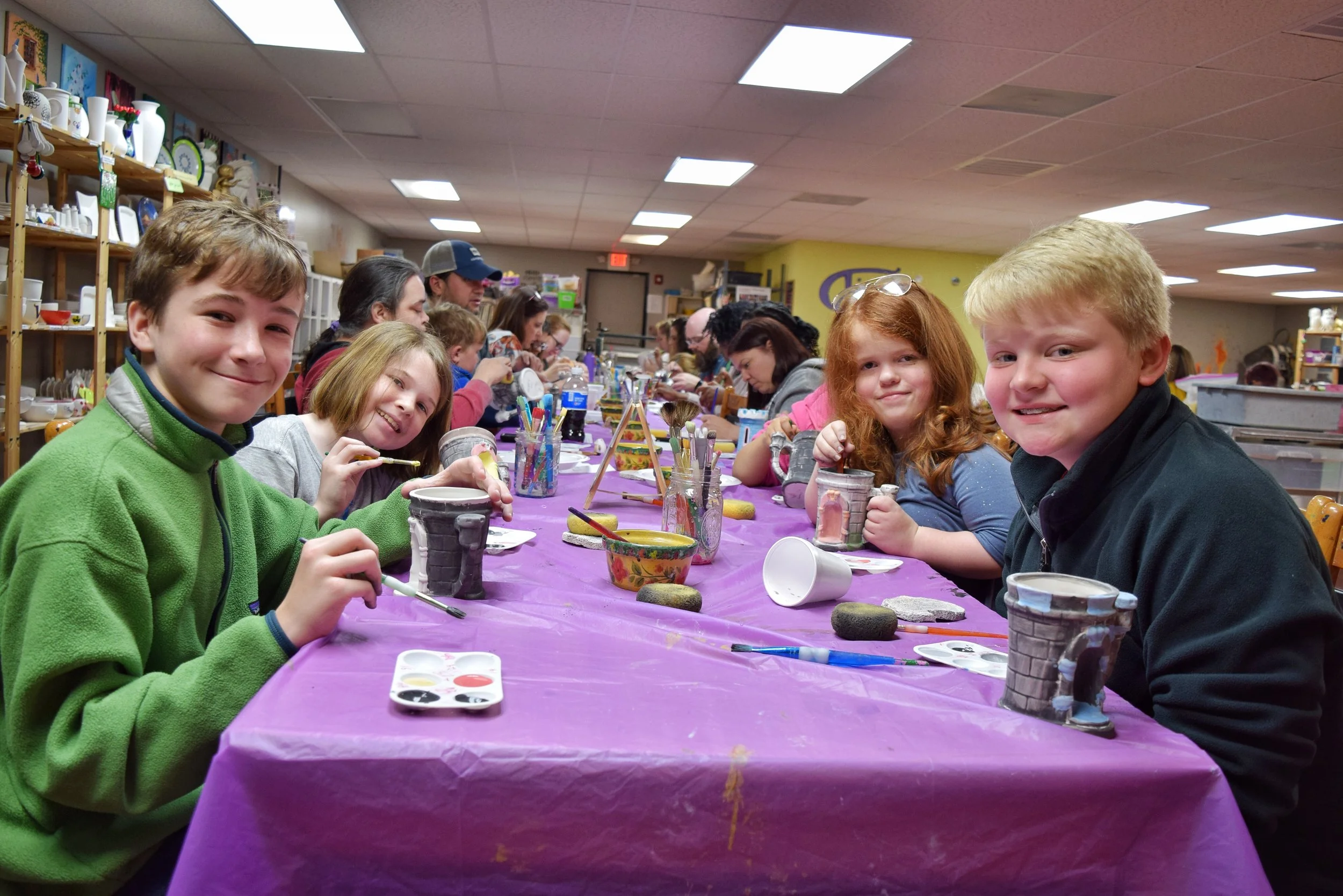 Children sitting at a long table covered with purple tablecloth, engaging in a pottery painting activity in a classroom or art studio. They are smiling and holding up painted mugs, with art supplies scattered across the table.