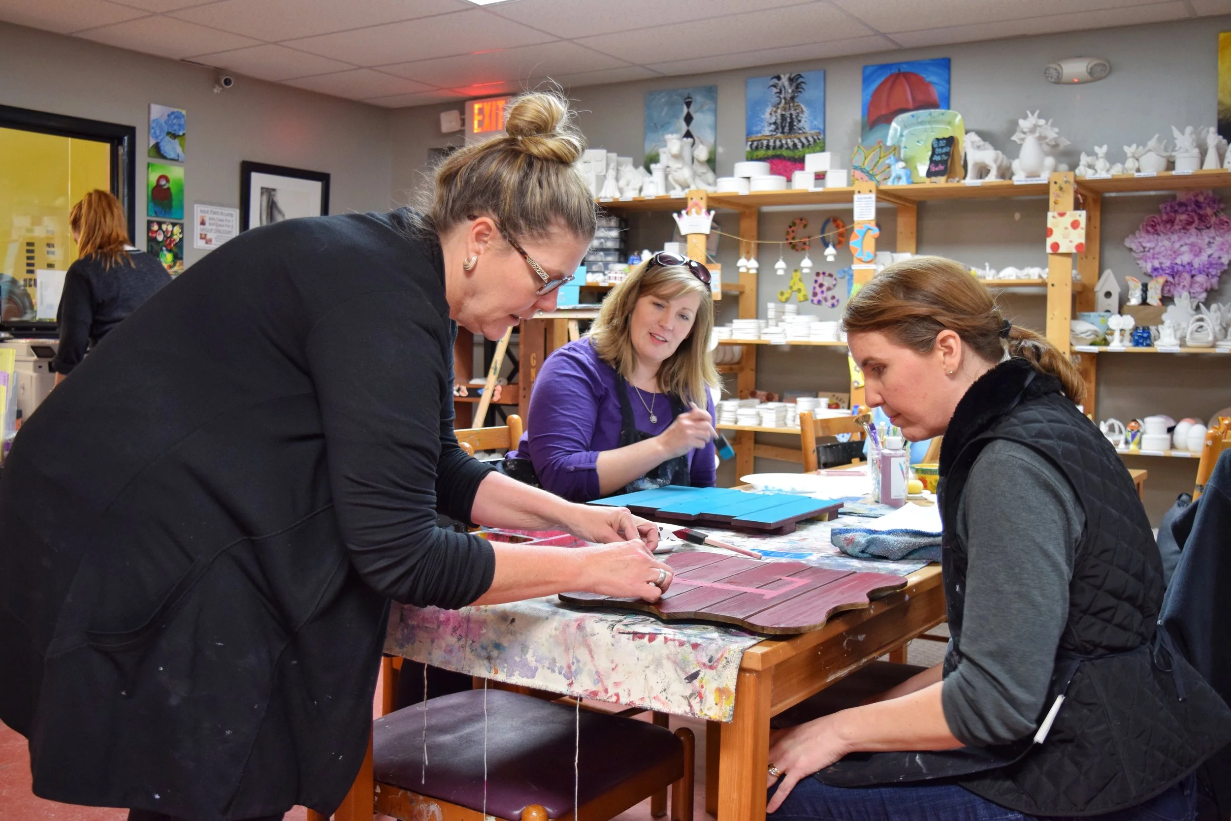 Three women in an art studio working on painted wood projects at a table. One woman is standing and leaning over, applying paint or glue to a wooden piece. The other two women are seated, one observing and the other working on her project.